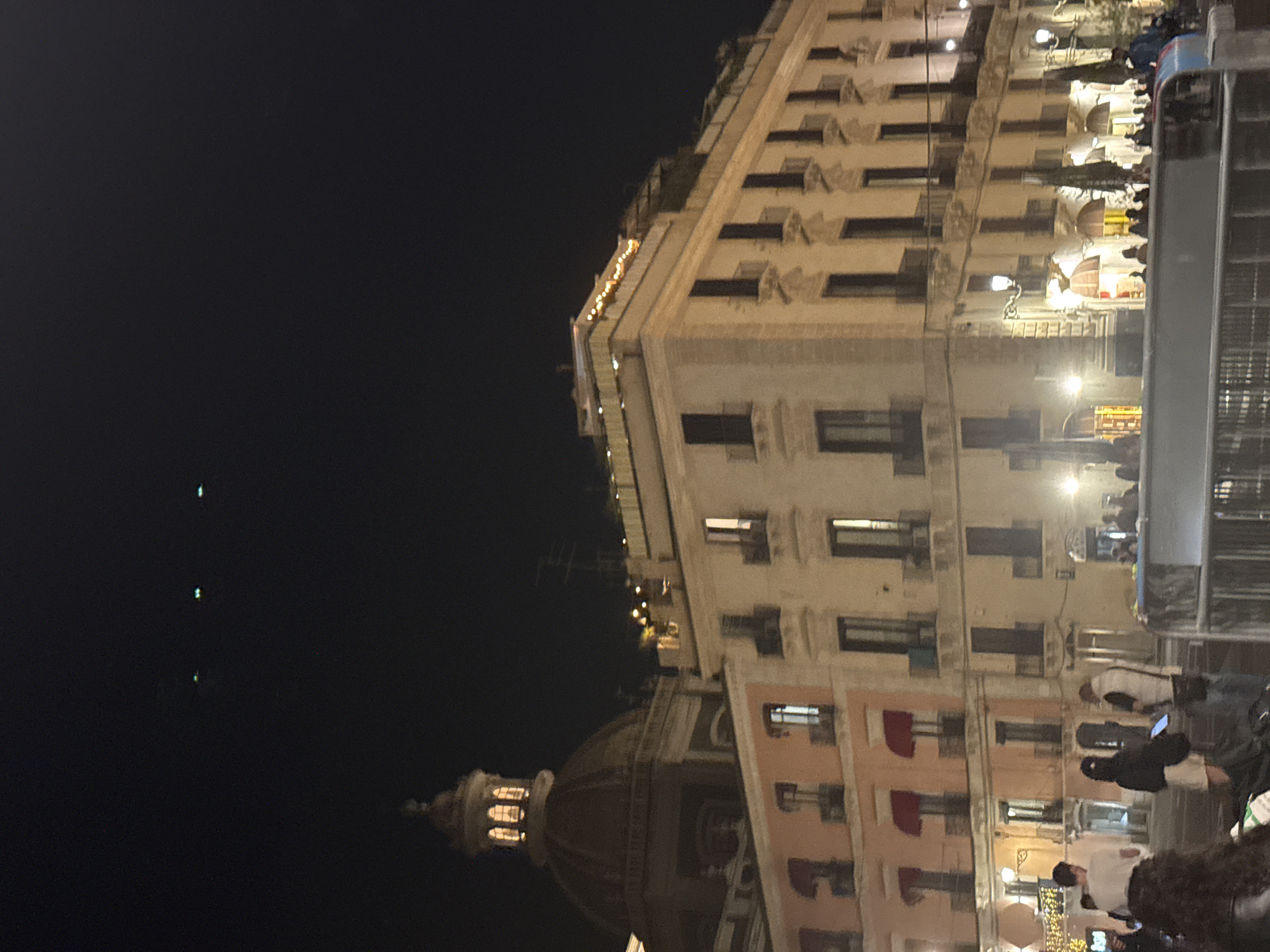 The image shows a nighttime view of a city street in Unknown, Unknown. A large building with multiple floors and windows dominates the right side of the image. The building is illuminated by streetlights and lights from within. A dome-shaped structure is visible on the left side, with its own set of lights. There are people walking and standing on the street, suggesting an active social environment. A vehicle, possibly a bus or trolley, is also present on the right side of the image. The sky is dark, with a few faint lights visible.
