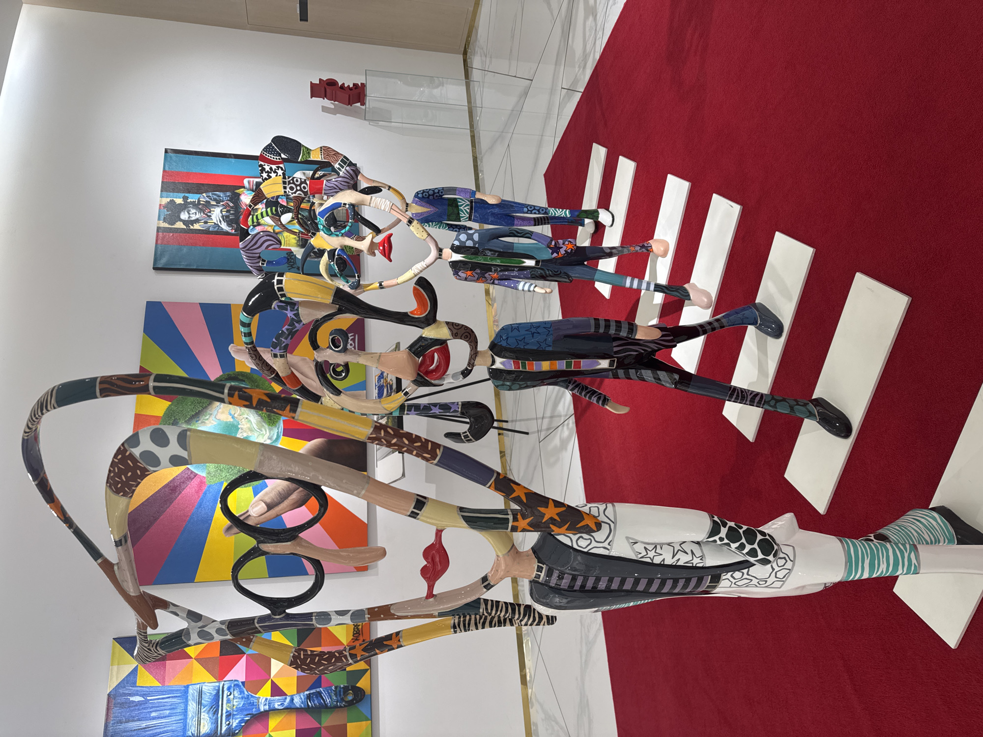 The image displays an art installation featuring colorful, stylized sculptures of figures walking across white rectangular pedestals arranged like a crosswalk on a red carpet. The sculptures have abstract, elongated heads with various patterns and colors, large glasses on one figure, and detailed clothing designs. In the background, vibrant abstract paintings adorn the white walls of what appears to be an art gallery or exhibition space. The floor is white and reflective. The scene is indoors, illuminated by overhead lighting, and exhibits a modern, artistic aesthetic.