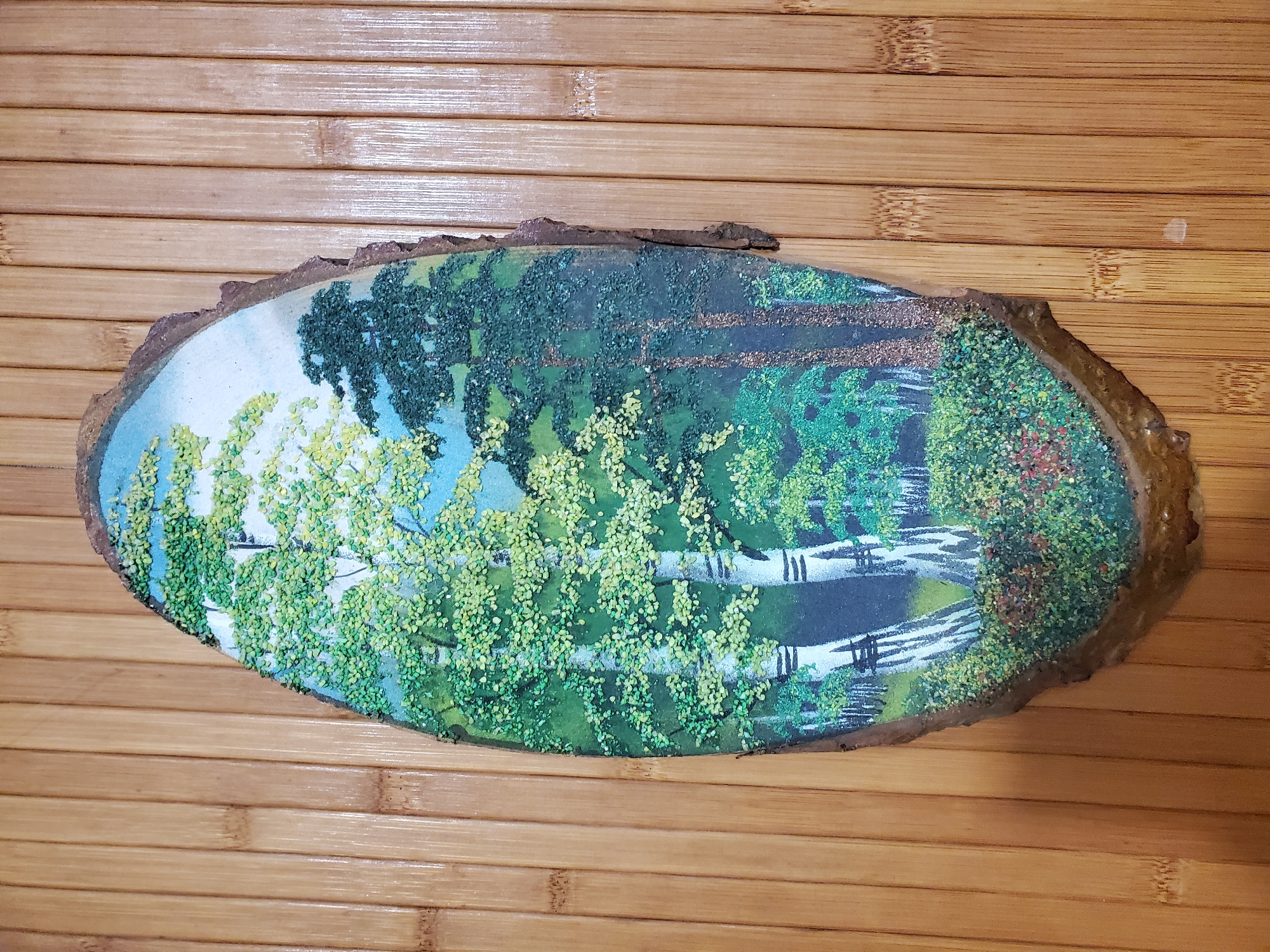 The image displays a piece of art on a piece of wood, likely a slice of a tree trunk, with a rough bark edge. The artwork itself is a painted forest scene with a textured application of small, colorful materials to represent foliage and ground cover. In the foreground, there are several birch trees with white bark and black markings, their leaves depicted with tiny green and yellow dots. To the right, a cluster of darker, needle-leaved trees stands, their forms rendered with denser, darker textured dots. The ground beneath the trees is covered in a thick layer of green, yellow, and reddish-brown textured dots, suggesting undergrowth and flowers. The background shows a lighter sky. The artwork is placed against a backdrop of vertically aligned bamboo slats, which have a natural, light brown color and visible nodes. There are no people or animals visible in the image. The location in Chornomorske, Ukraine, is not discernible from the visual content of the image.