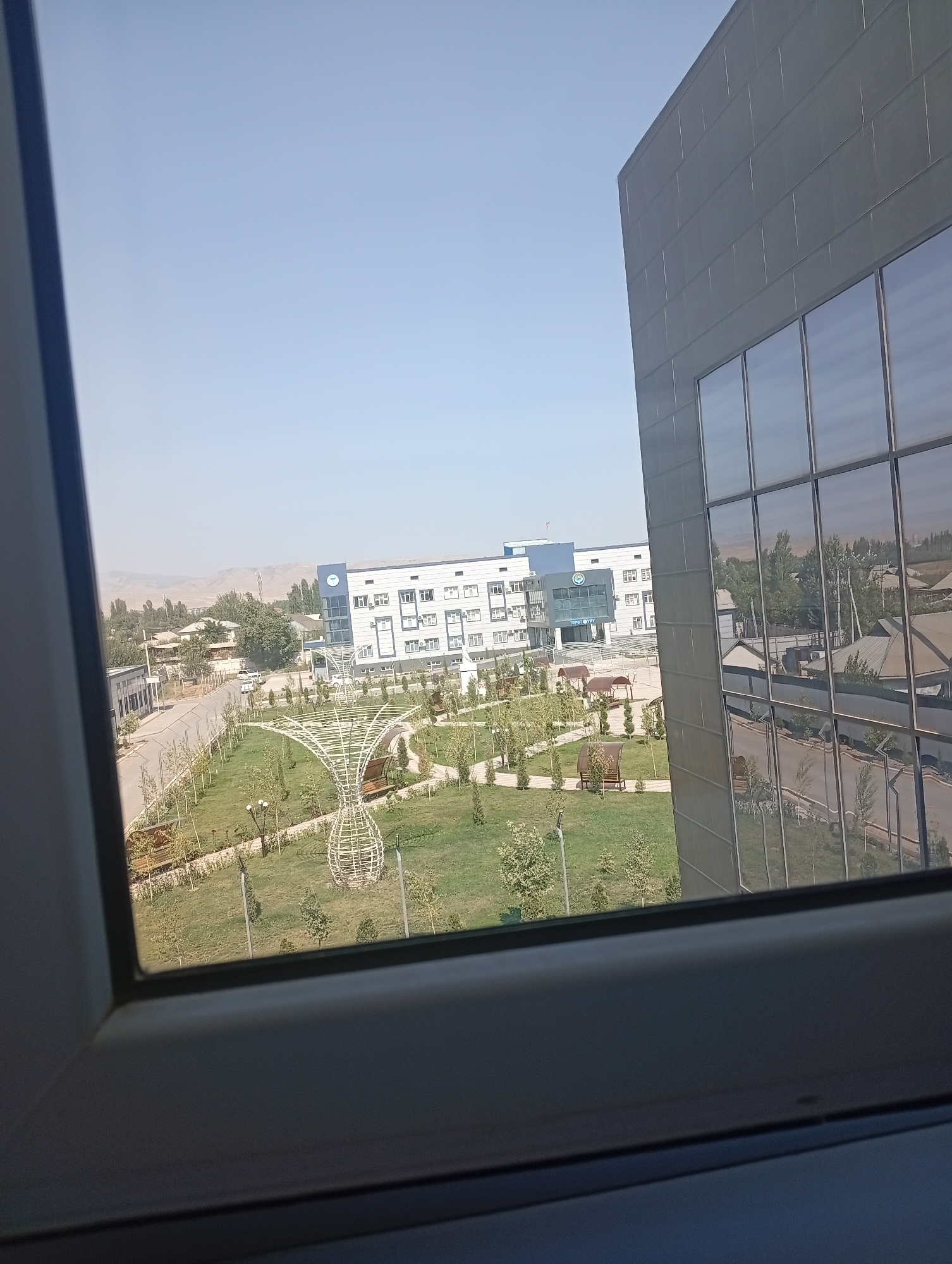 A high-angle, medium shot captures a view through a window frame of a modern building and a surrounding park area in Türk-Abad, Kyrgyzstan. The foreground is dominated by the dark gray window frame, obscuring the bottom portion of the image. To the right, the reflective glass facade of a contemporary, multi-story building rises, its dark gray panels and reflective windows dominating the right half of the frame. The windows offer distorted reflections of the park and surrounding buildings.
The middle ground reveals a well-maintained park with a neatly mowed green lawn, scattered young trees, and paved pathways. A distinctive white, wireframe sculpture, resembling an abstract vase or flower, stands prominently in the park. Benches are visible along the pathways. Beyond the park, a road runs horizontally, with a few cars visible. On the left side of the road, a low-rise building with white walls and a blue roof is situated. Further in the distance, more buildings, trees, and hilly terrain can be seen under a clear, pale blue sky. The overall impression is one of a modern institutional or residential area with recreational green spaces.