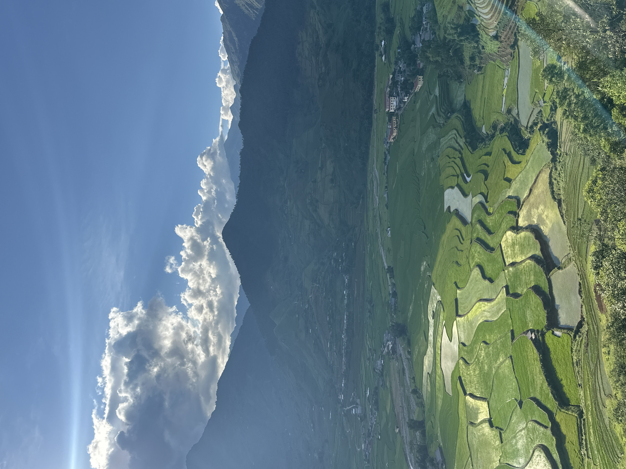 This image presents a panoramic view of a mountainous, agricultural valley in Tú Lệ, Vietnam, under a partially cloudy sky. The upper portion depicts a clear blue sky interspersed with both brilliant white cumulus clouds and darker grey clouds, some of which are illuminated from behind by sunlight. A prominent sunbeam or lens flare is visible in the upper left, emanating from behind the clouds.
The mid-ground is dominated by multiple layers of dark, verdant mountains, their peaks often silhouetted against the brighter sky, extending across the entire width of the frame. Shadows are cast heavily on their slopes.
The valley floor, which occupies the lower two-thirds of the image, is characterized by extensive terraced rice paddies. These paddies are irregular in shape and vary in color from bright lime green to deeper emerald, with many sections submerged in water that reflects the sky and sunlight, creating luminous, silver-toned patches. A winding river or stream is visible traversing the valley floor, alongside which are several clusters of buildings forming a village or settlement. These structures, primarily light-colored with darker roofs, are interspersed among the rice fields, with a more concentrated group featuring yellow-walled buildings visible towards the mid-right. Roads or paths are discernible connecting parts of the settlement. Dense green foliage and trees line the lower slopes of the mountains and are present in the immediate foreground, particularly on the right, partially obscuring the lower-right corner of the view, where another lens flare is present. No people or animals are visible. The lighting suggests either morning or late afternoon.