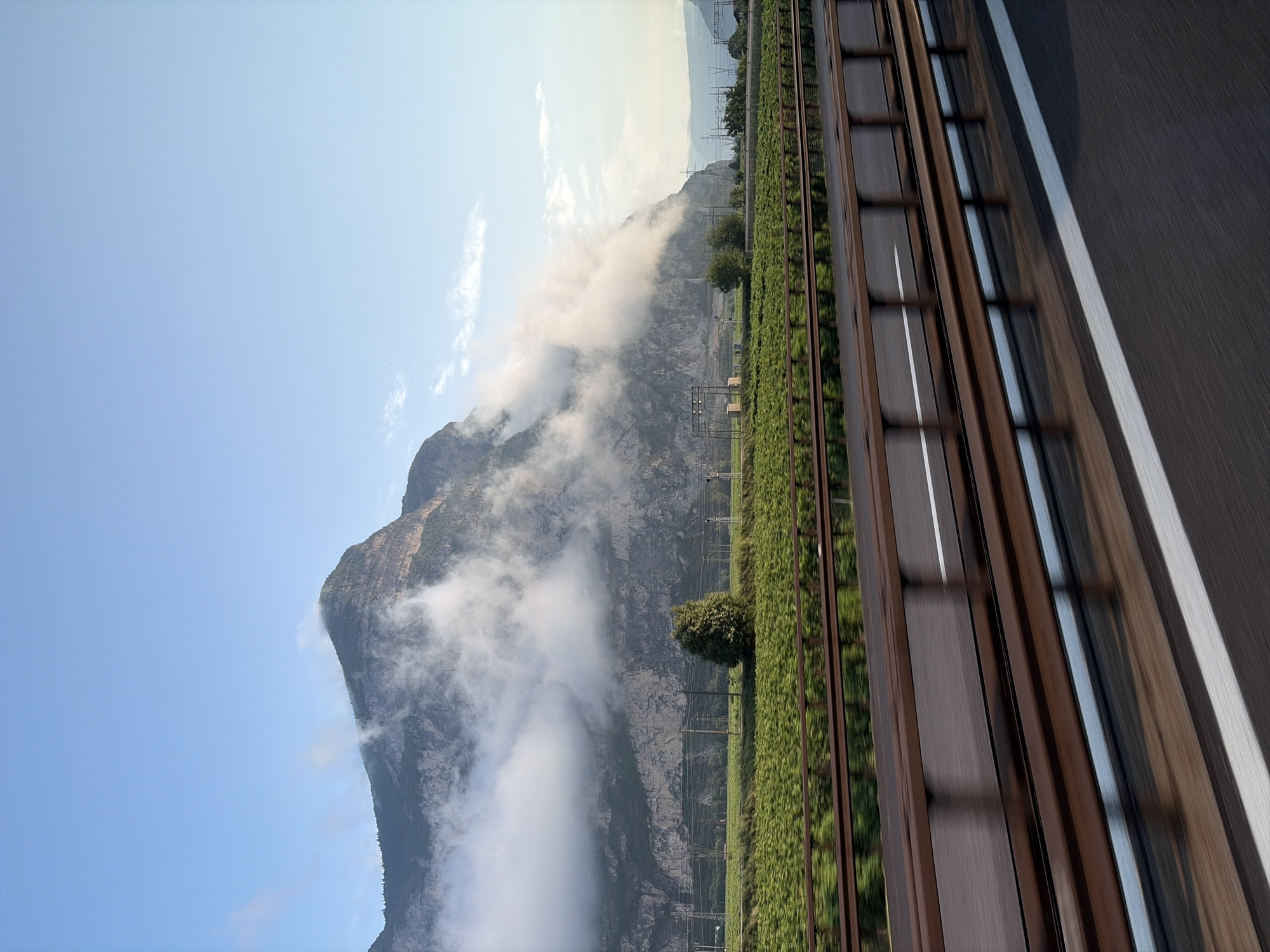 The image shows a mountain range in Neumarkt - Egna, Italy, partially shrouded in clouds. In the foreground, there is a highway with guardrails, indicating motion blur consistent with travel. A field of green crops stretches between the highway and the mountain base, with power lines and a few trees visible. The sky is clear blue with some wispy clouds. The scene captures a scenic view from a moving vehicle.