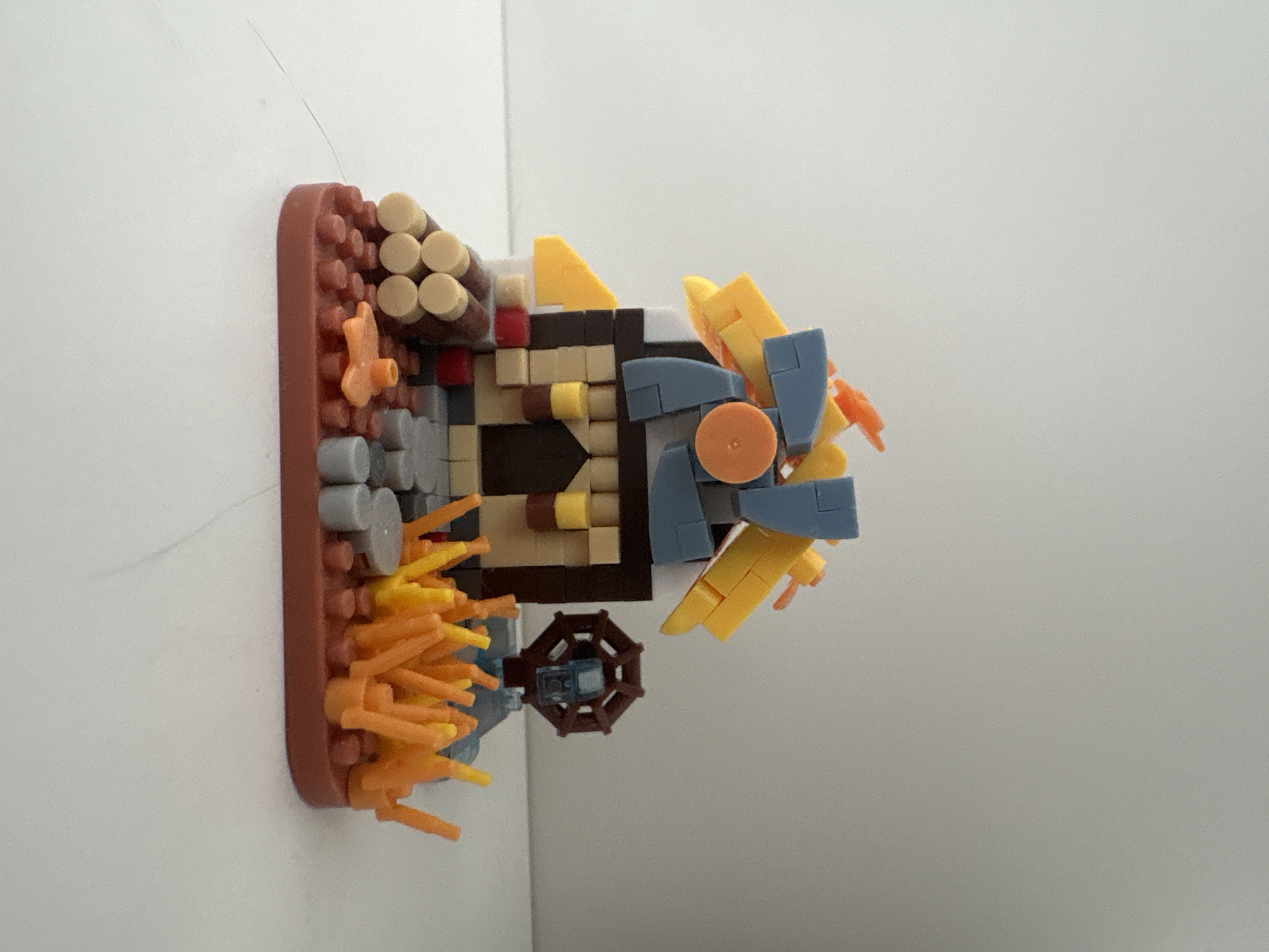 This is a medium-shot photograph of a small, colorful building made of interlocking plastic bricks, resembling a simplified windmill or barn structure. The structure has a dark brown base with tan, white, and dark brown bricks forming the walls and a dark doorway. The roof is a complex arrangement of yellow, orange, and blue angled bricks, with some orange flame-like pieces at the very top. To the left of the structure, there are several brown and light tan cylindrical pieces stacked horizontally, representing logs. In front of the structure and to the right, there are clusters of bright orange and yellow thin, upright pieces that resemble flames or tall grass. A small, brown, octagonal wheel with blue accents is visible to the right of the building. The entire structure sits on a flat, brown, brick-shaped base. The background is a plain, light gray, out-of-focus wall, and the foreground is a white surface. There is no indication of the city of Kyiv, Ukraine, or any other specific location. The image contains no people or discernible actions.