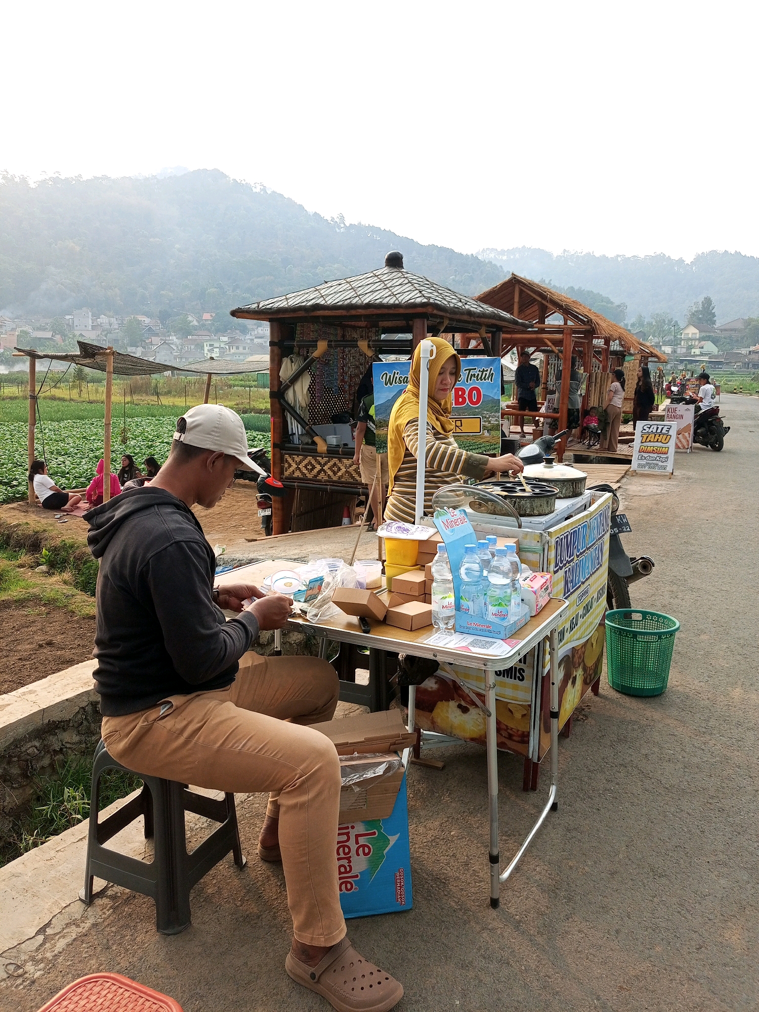 A food stall is set up on the side of a paved road, with a rural landscape of green fields and hills in the background, likely in the city of Batu, Indonesia, known for its agricultural tourism. A woman wearing a yellow hijab and striped shirt is operating the stall, cooking what appears to be mini pancakes in a specialized pan. She is interacting with a customer, a man in a dark hoodie and tan pants, who is seated on a stool in the foreground, looking down at something he is holding in his hands. The stall itself is a mobile cart adorned with colorful signage advertising food items like "Sate Tahu" and "Dimsum". Various items are displayed on and around the stall, including boxes of food, bottles of water, and a green basket. In the background, other people are visible, some sitting near a traditional-looking structure, and a motorbike is parked nearby. The overall scene depicts a typical day at a roadside food vendor in a scenic, semi-urban or rural area.