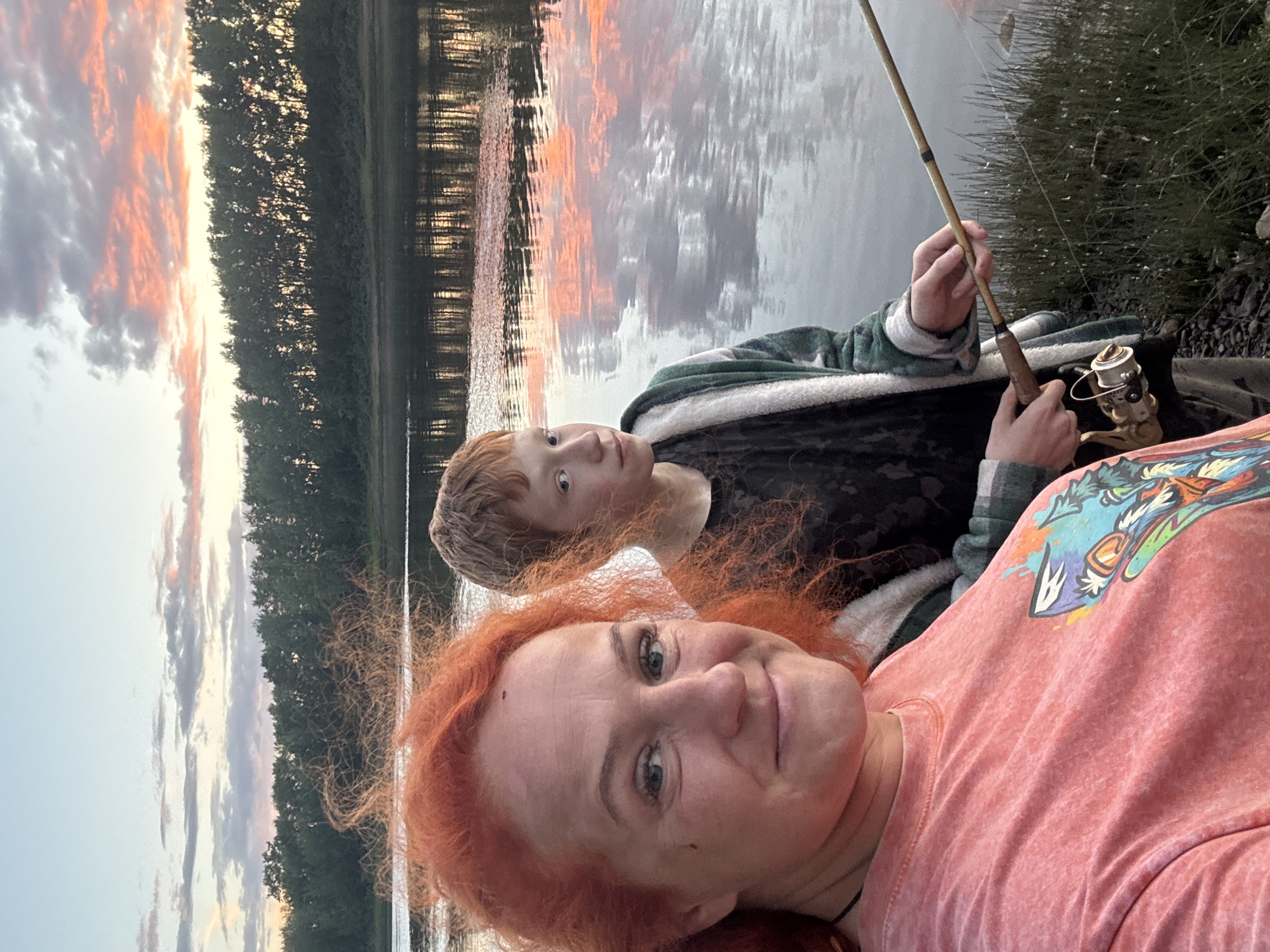 The image shows a close-up selfie of two people with a natural landscape in the background. In the foreground, a woman with vibrant red hair is looking towards the camera with a slight smile. She is wearing a pink t-shirt with a colorful graphic print. Behind her, a younger individual with red hair is also looking towards the camera. This person is wearing a green and white checkered robe or loungewear. They are holding a fishing rod, suggesting the activity of fishing.
The background depicts a serene waterscape, likely a lake or river, reflecting the sky and a dense line of trees along the distant bank. The sky is filled with dramatic clouds tinged with orange and pink, indicating either sunrise or sunset. The water has a textured surface with ripples, further reflecting the sky and vegetation. The overall mood is peaceful and relaxed, with the warm colors of the sky creating a picturesque setting. The image appears to have been taken outdoors, in a natural environment, possibly during an evening or early morning fishing excursion. There is no visible text in the image.