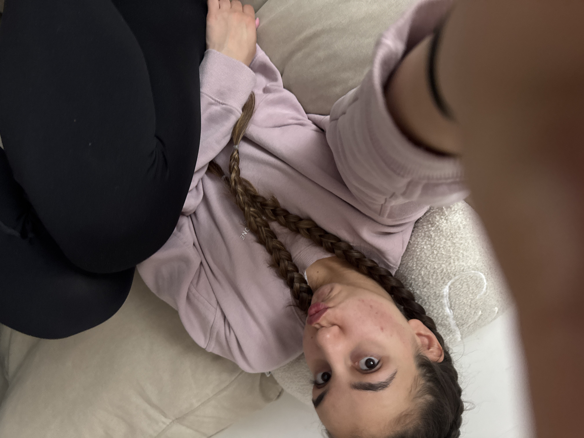 A young woman is lying on a light beige-colored couch, wearing black leggings and a light pink sweatshirt with two braids in her hair. The image appears to be taken in Sofia, Bulgaria. She is holding the camera in front of her face, slightly obscuring the view. The background consists of the couch.