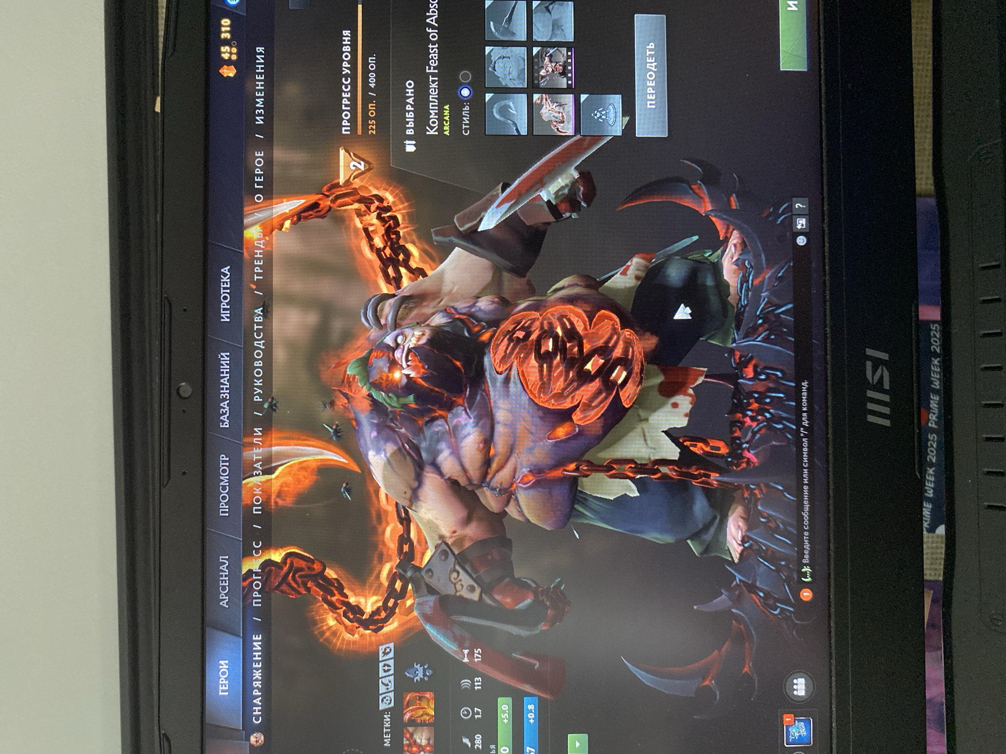 The image shows a close-up view of a laptop screen displaying the game Dota 2. The game's interface is visible, with menus and character art. A large, monstrous character in fiery chains is prominently featured in the center of the screen. The laptop itself appears to be placed on a surface, possibly a desk, with a blurred background that offers no discernible context for its location. There is no indication of the city of Łódź, Poland, or any other geographical location.