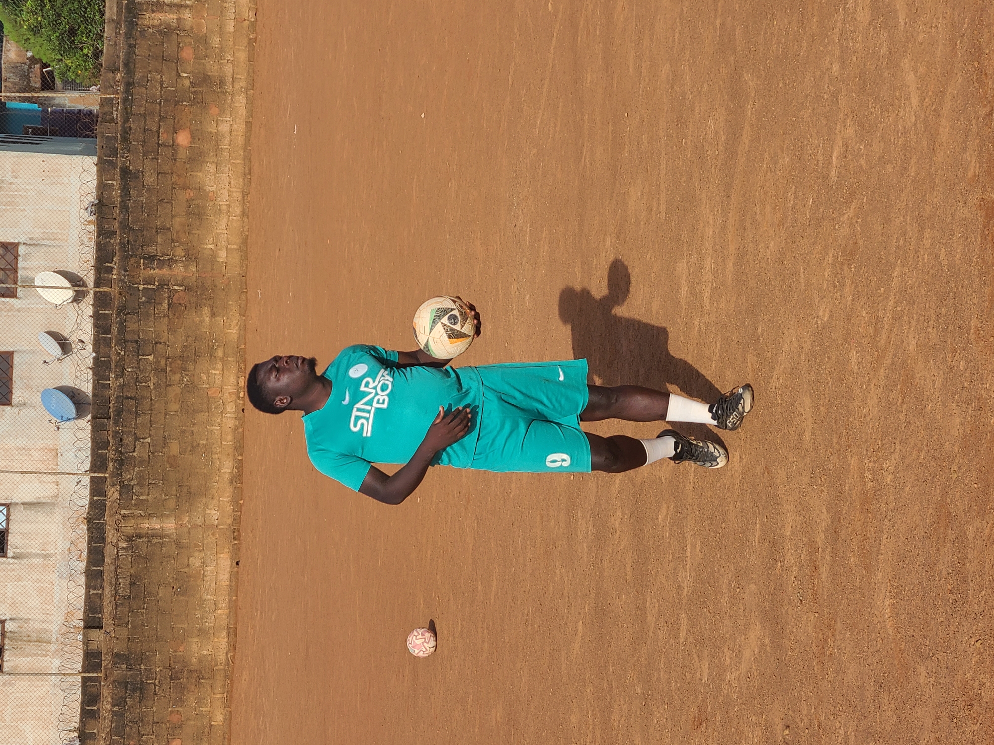 A young Black man is standing on a dirt soccer field in Barakin Lerawi, Nigeria, holding a soccer ball in his right hand. He is wearing a turquoise t-shirt with the white text "STAR BOY" and matching turquoise shorts with the number "9" printed in white on the left thigh. His socks are white, and he is wearing black and white soccer cleats. The field is made of packed reddish-brown dirt. In the background, there is a low brick wall topped with a chain-link fence and barbed wire. Behind the fence, a light-colored building with barred windows and two satellite dishes is visible. Another smaller pink soccer ball is on the field to the left of the man. The lighting suggests it is daytime, casting a shadow to the man's left.