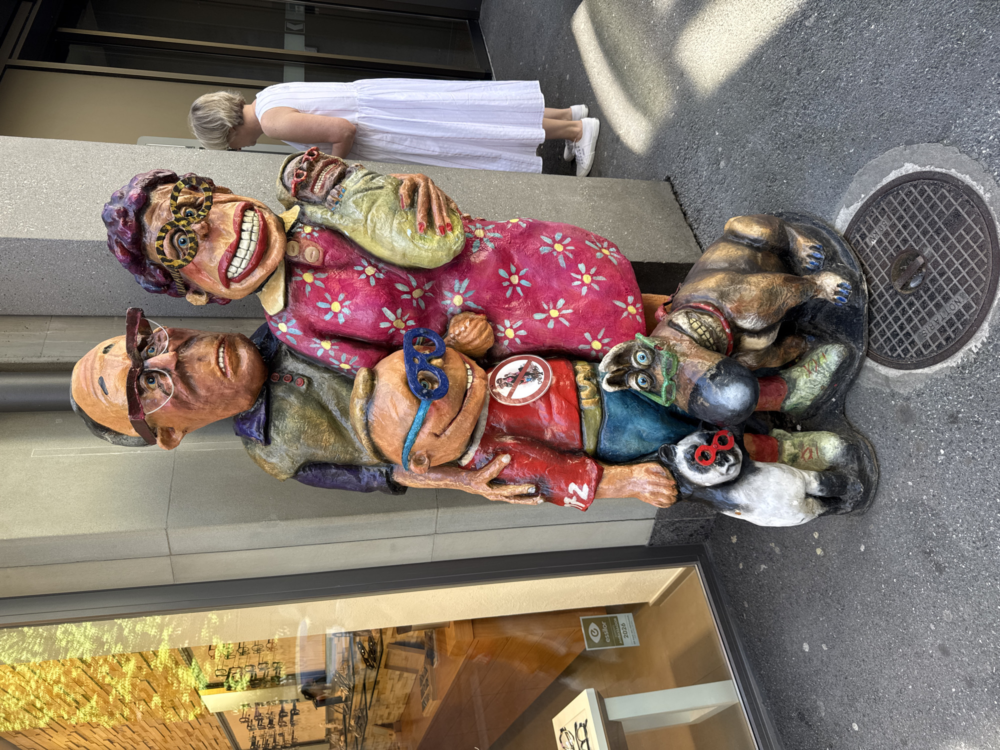A vertical, eye-level shot features a colorful, life-sized sculpture of a caricatured family with a dog, placed on a sidewalk in what appears to be a European city. The sculpture is positioned to the right of a building with a large glass storefront, reflecting trees and brickwork. To the left, a woman in a white dress and white sneakers bends over the sculpture, her blonde hair visible.
The sculpture depicts a woman with exaggerated features, wearing a pink floral dress and large, circular glasses with a leopard print frame. She clutches a green bag and has a wide, toothy grin. Below her, a man with glasses and a surprised expression is partially visible. Other figures and animals, including a dog and what looks like a panda, are also part of the sculpture, all rendered with exaggerated and humorous features. Some figures have red accents, like fingernails and glasses. A circular sticker with a motorcycle symbol and a red circle with a slash through it is attached to the woman's dress.
The scene is outdoors, under daylight, suggesting a pleasant weather day. A manhole cover with text is visible on the asphalt in the background. The storefront glass displays eyeglasses and promotional materials. A visible sign on the storefront reads "essilor" with "MALAKO" and "2026" below it. The overall impression is a lighthearted and whimsical street art display.