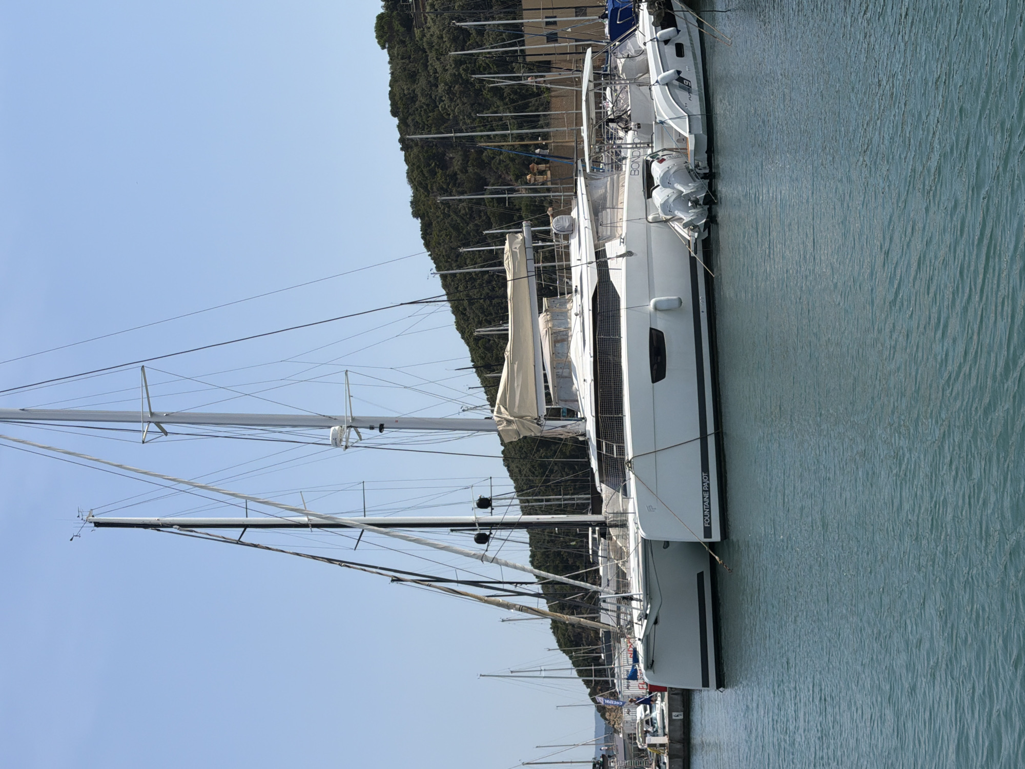 A wide shot shows a fleet of sailboats moored in a harbor under a clear blue sky. In the foreground, two large white catamarans are docked side-by-side. The catamaran on the right has the name "FOUNTAINE PAJOT" emblazoned on its hull. A smaller white boat with twin outboard motors is attached to the stern of the catamaran on the right. Behind the catamarans, numerous other sailboats and masts are visible, extending towards a densely wooded hillside. The water is a deep blue-green, reflecting the bright sunlight. The scene appears to be in the town of Punta Ala, Italy, known for its marina and scenic coastal landscape.