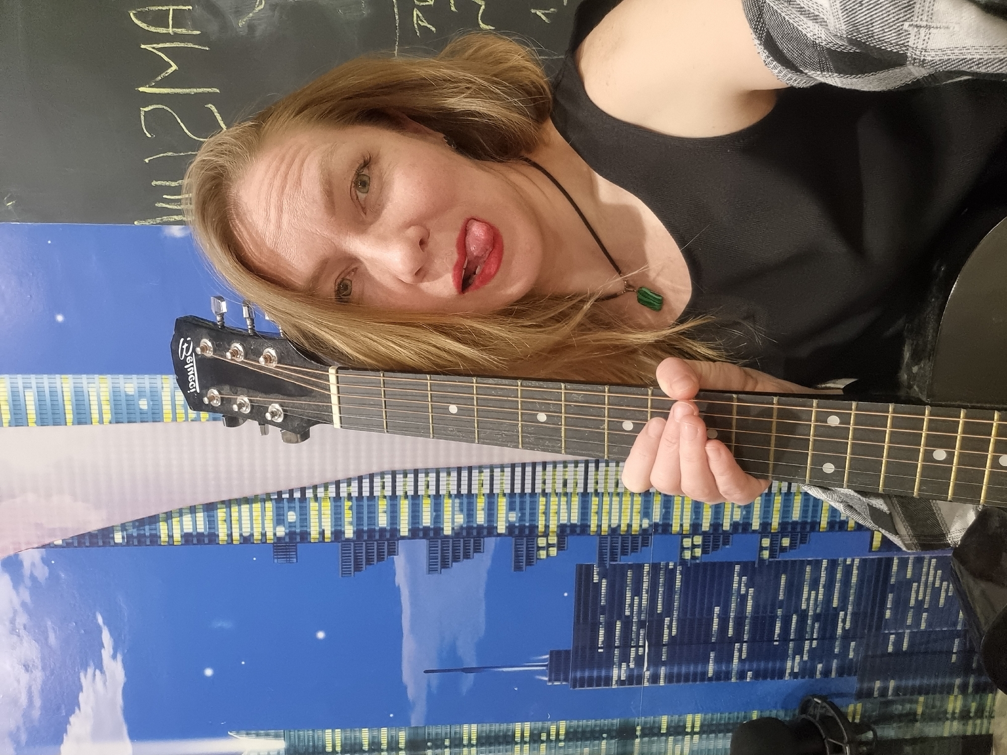 A woman is holding a guitar while standing in front of a poster with a city background. The woman has blonde hair, is wearing a black tank top, a plaid shirt, and a green pendant necklace. She is sticking her tongue out with red lipstick on her lips. The head of the guitar is visible on the left. The city poster behind her has a blue sky with white clouds and buildings. Above the poster, a chalkboard with writing is partially visible. The location is not identifiable as Chaikovsky, Russia.