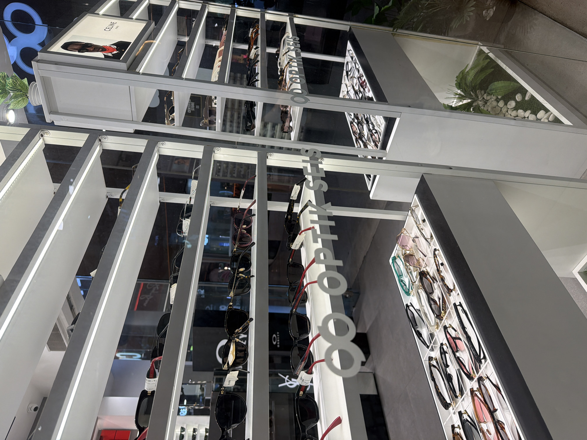 The image shows a display of eyeglasses and sunglasses inside a store in Kuta, Indonesia. The display consists of a white, multi-tiered shelving unit with rows of glasses hanging on hooks. The brand "CELINE" is prominently displayed on one of the shelves. A decorative plant is visible at the top of the unit. A white sign that says “Seis Optik Co.” is mounted on the side of the shelving unit.