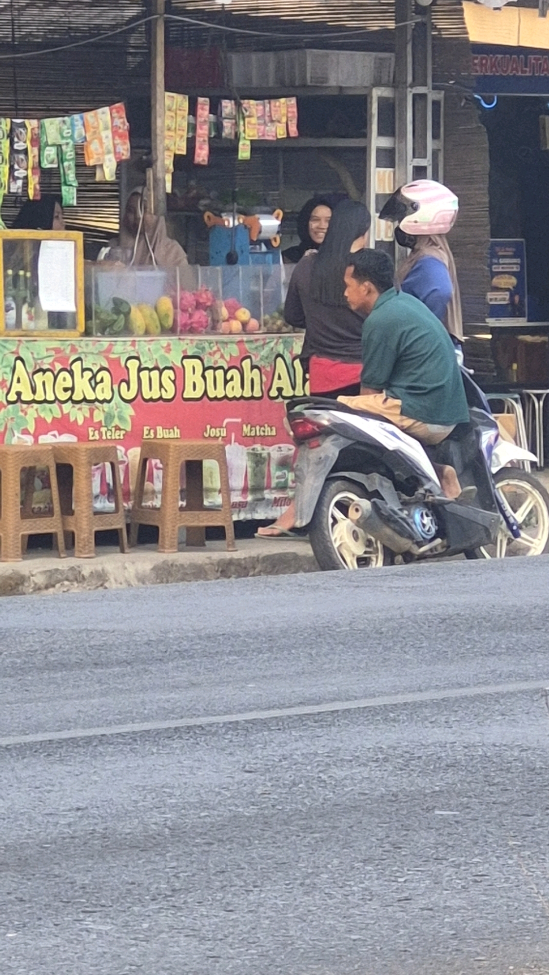 The image depicts a street scene in Putak, Indonesia, likely in front of a roadside food stall. The stall features a red banner with yellow lettering that reads "Aneka Jus Buah Al" (Various Fruit Juices). Below the banner are several plastic stools and an array of fruits, including dragon fruit, mangoes, and possibly other tropical varieties. Behind the stall, several people are visible. Two women, one wearing a black hijab and the other with a pink and white helmet, are standing near the stall. A man in a green shirt and light-colored shorts is sitting on a motorcycle, parked alongside the stall. The motorcycle is a dark color with some white accents and a silver exhaust pipe. The background shows a building with awnings and more signage, some of which is partially obscured. The foreground is dominated by a paved road with a white line marking.