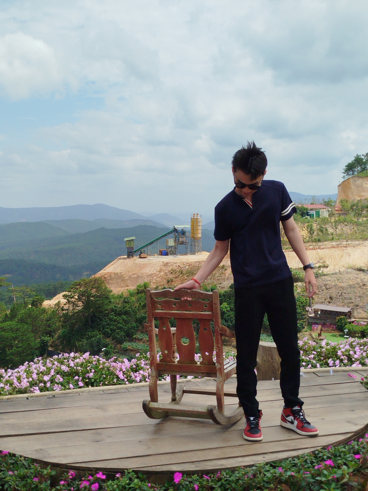 An Asian man, wearing a dark blue polo shirt with white striped sleeves, black pants, sunglasses, and red and black sneakers, stands on a wooden platform. He is leaning his right hand on the armrest of a wooden rocking chair and looking down. The background features a lush green mountainous landscape with a quarry or construction site in the mid-ground, including machinery and stacked materials. Pink flowers are visible in the foreground along the edge of the platform. The sky is partly cloudy. The scene appears to be an outdoor viewpoint, possibly in Xuân Thành, Vietnam, given the mountainous terrain and the presence of pink flowers.