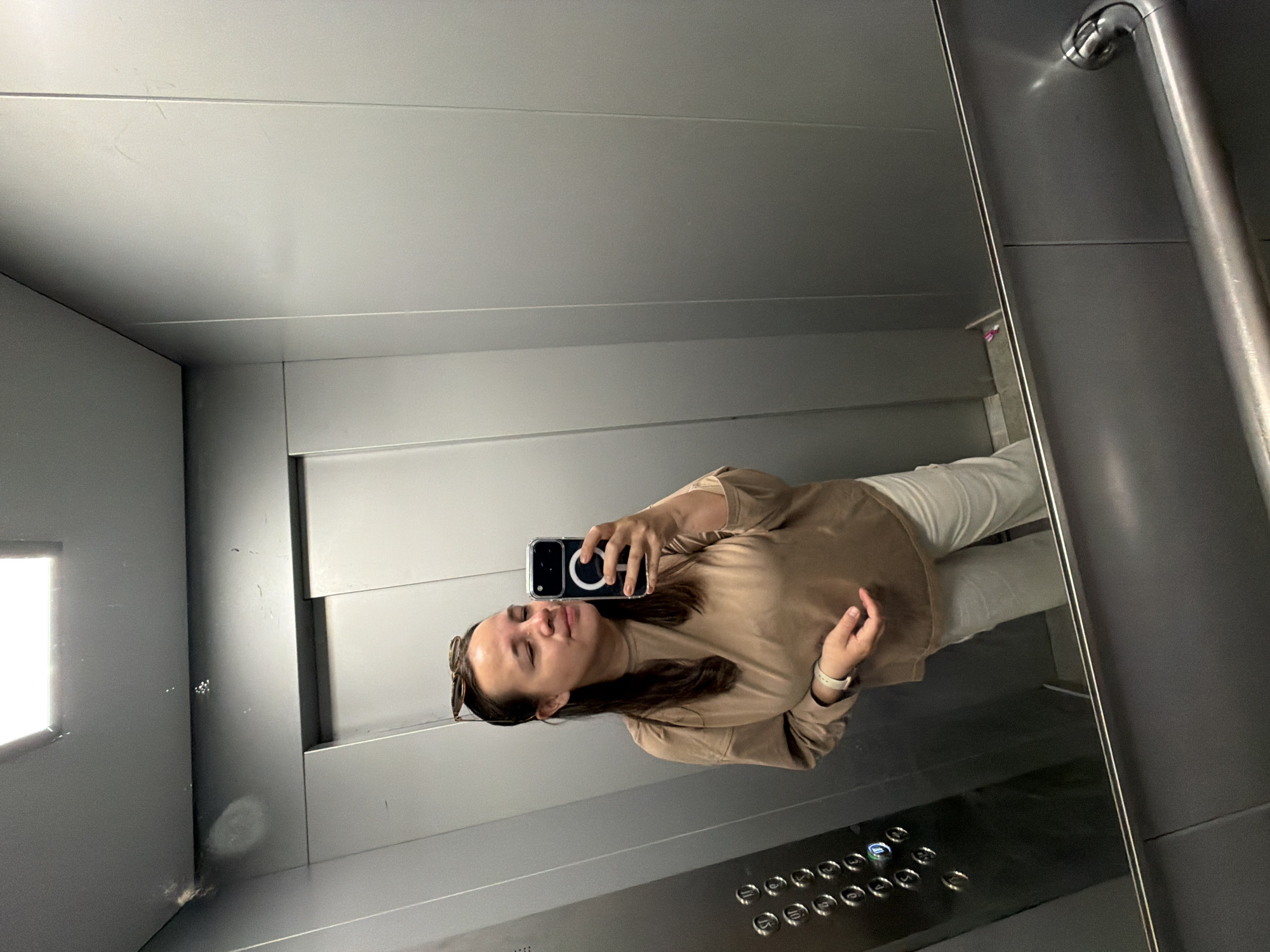 The image shows a person taking a selfie in what appears to be an elevator. The individual is wearing a light brown shirt and white pants. They are holding a smartphone. The elevator has metal walls and a panel with buttons. Based on metadata, the location is Chaikovsky, Russia.
