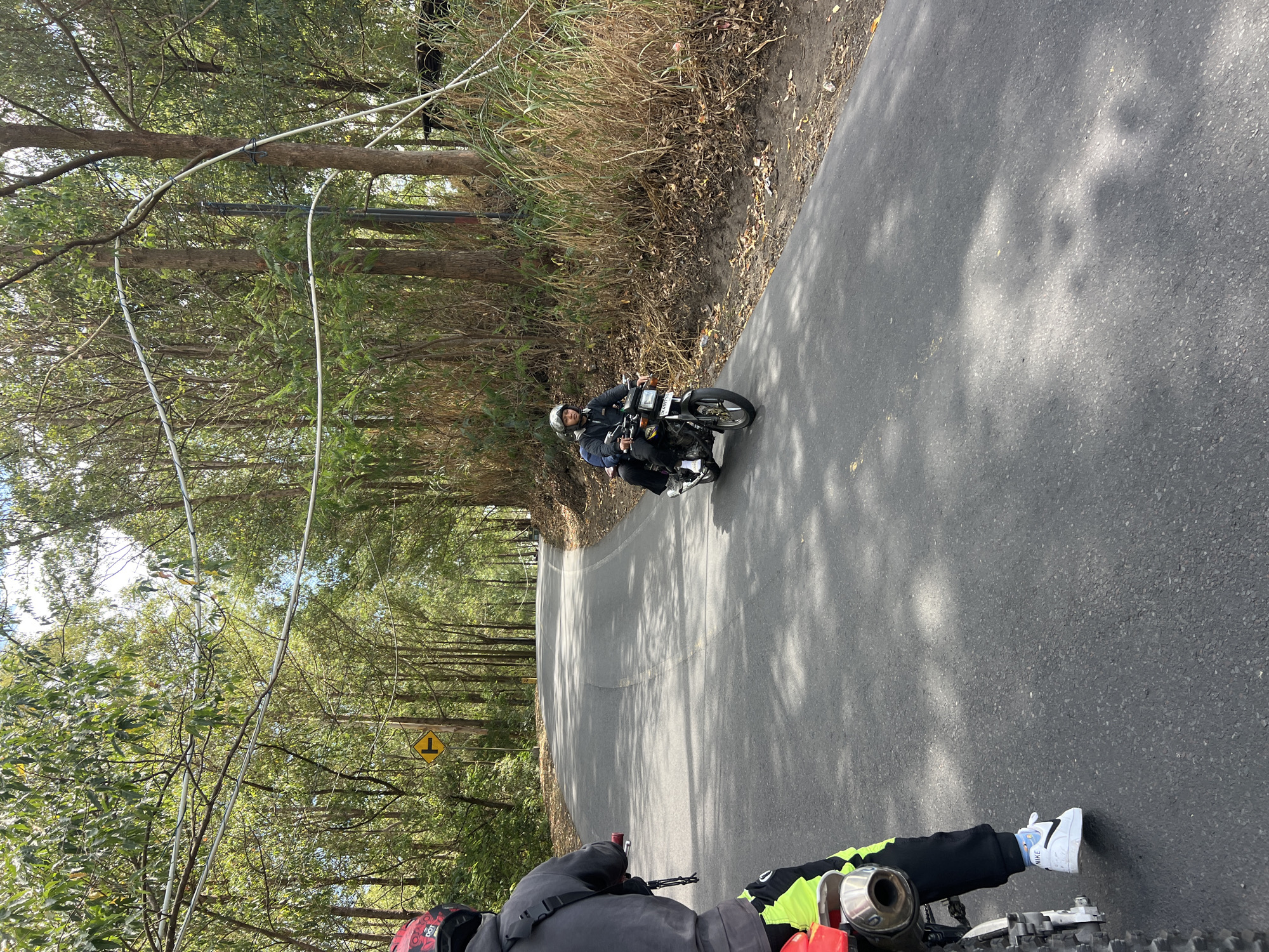The image shows two individuals riding motorcycles on a paved road in a rural or semi-rural area, likely in Kintamani, Indonesia, given the vegetation and terrain. The road is asphalt and appears to be a two-lane road winding through a wooded area with tall, slender trees and dense foliage. Overhead, electrical wires are strung between poles, arching across the road.
In the foreground, the back of a person on a dirt bike is visible, facing away from the camera. This person is wearing a dark gray hoodie, black pants with bright neon yellow accents, and white Nike sneakers. A black backpack strap is visible over their left shoulder.
Further down the road, another motorcycle is approaching, carrying two people. The rider is wearing a helmet and a dark jacket, and the passenger is wearing a helmet and a dark top. The motorcycle is a dark color with a visible license plate.
A yellow directional sign with a black arrow pointing right is visible on the left side of the road in the mid-ground. The ground on the sides of the road is covered with dry leaves, grass, and dirt. Sunlight filters through the trees, casting dappled shadows on the road surface. The overall impression is a scenic, natural environment with human activity.
