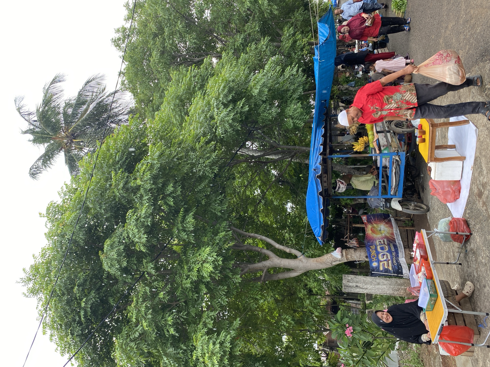 In an outdoor market scene, a woman wearing a black hijab and traditional attire sits at a small yellow table laden with food containers. She is smiling. The table is set up on a wooden plank, and red plastic bags are placed around it. Behind her, a concrete pillar stands next to a large tree with dense green foliage. A banner with the text "EDGE PUNCH" is visible. Further to the right, a blue awning covers a stall with a motorcycle parked nearby. A man in a red patterned shirt and dark pants is walking away from the stall, carrying a large plastic bag. In the background, other people are present, and more trees and greenery dominate the landscape, suggesting a tropical or subtropical environment. The sky is overcast.