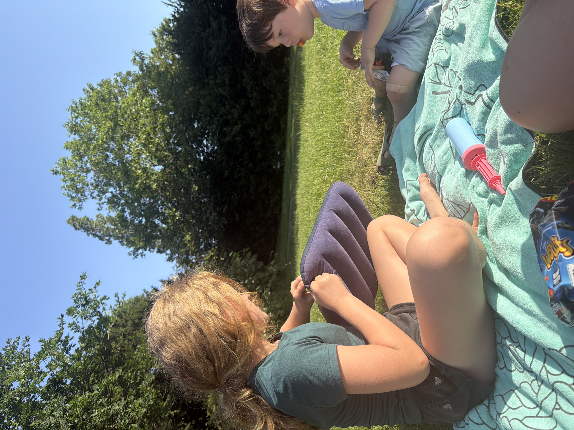 Outdoors on a sunny day in what appears to be a park, a young girl with blonde hair pulled back in a ponytail lies on the grass with a blue inflatable pillow. She is wearing a dark green shirt and dark shorts. A boy with brown hair wearing blue clothing stands nearby, looking down at something on the grass. A blue and red hand pump rests on a light blue patterned blanket. A package of Walkers crisps is also on the blanket. Green trees and a blue sky are visible in the background. The location is likely a park in London, United Kingdom, given the presence of Walkers crisps.