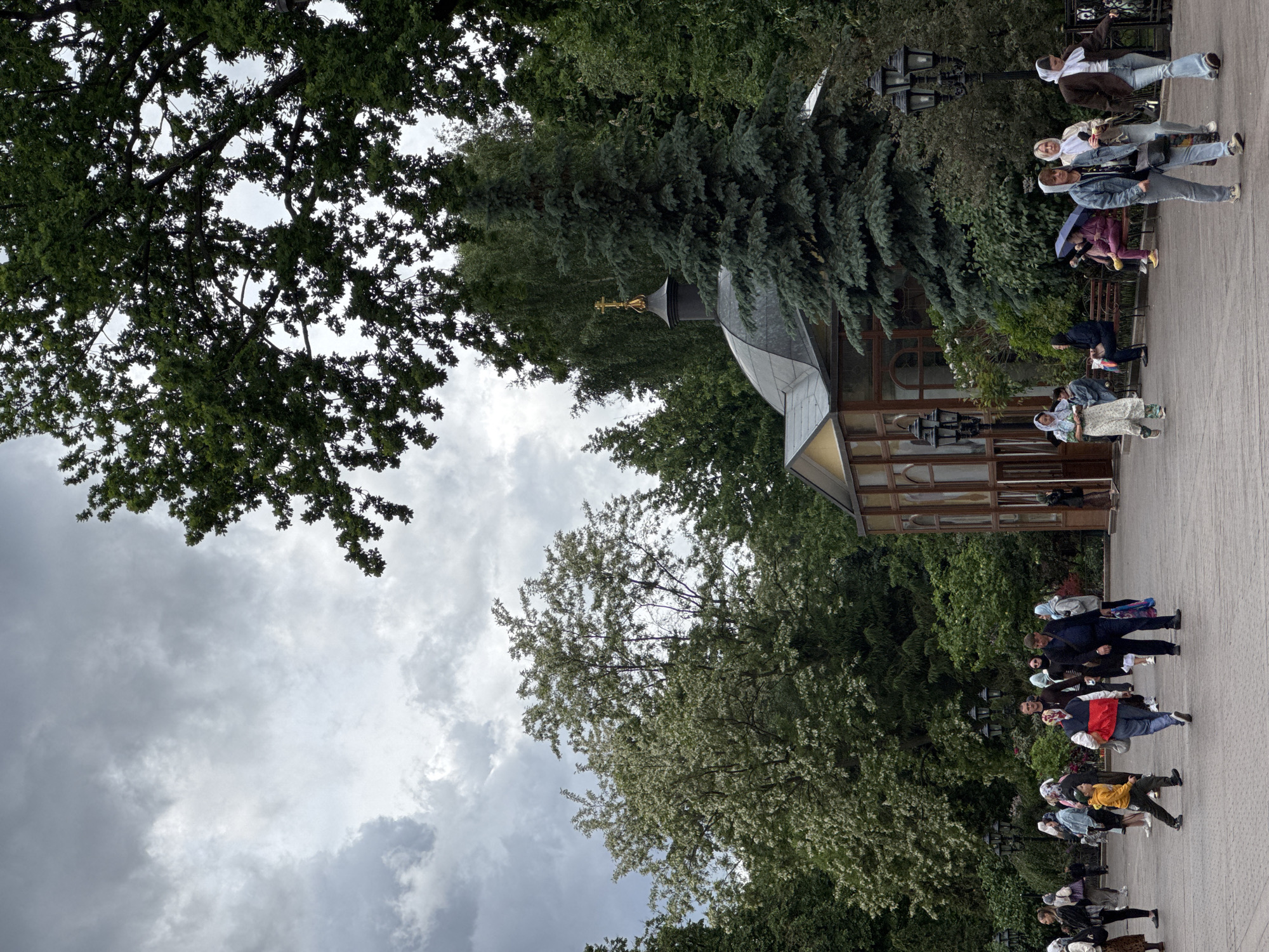 The image depicts a group of people walking along a paved walkway near a building with a distinctive roof and cross, likely a chapel or small church. The location appears to be Zaryadye Park in Moscow, Russia. The weather seems overcast, with a cloudy sky visible through the trees. People are casually dressed, with some individuals wearing headscarves, suggesting a possible religious significance. The presence of trees and greenery indicates a park or garden setting. The photograph shows a common urban scene with elements of religious architecture and public gathering space.