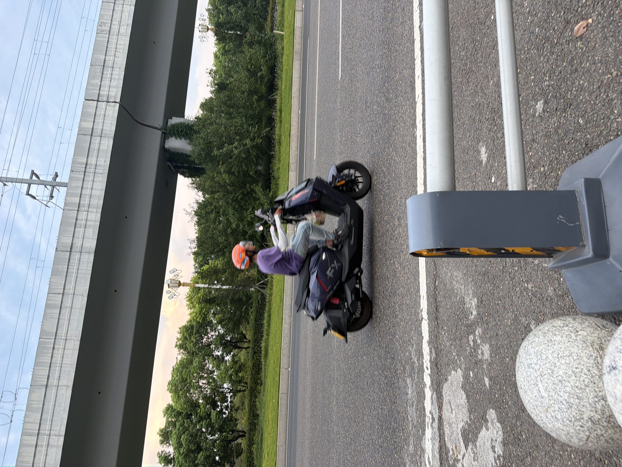 A person wearing an orange helmet and a purple shirt rides a dark-colored electric scooter down a paved road. The scooter has a passenger seat and a front fairing. The rider is wearing blue jeans and white gloves, and their left arm is extended. The road has white lane markings and is bordered by a grassy median and trees. Overhead, a concrete overpass runs across the image, supported by columns. The sky is overcast with hints of pink and yellow from what appears to be a sunset. In the foreground, a grey, textured traffic barrier and large round stones are visible. The scene appears to be in the city of Jiangjin, China, given the context provided.