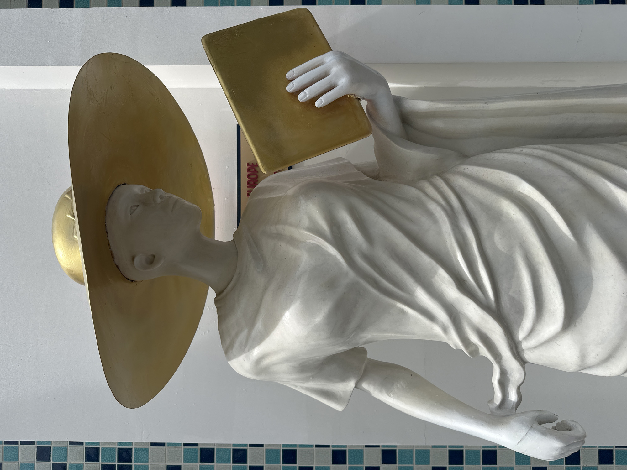 A statue of a figure wearing a golden hat and holding a golden book is presented. The statue is white and appears to be draped in flowing robes. It is positioned against a white wall with a band of blue and white mosaic tiles running along the bottom. Based on the location indicated, this statue is likely situated in or near Bac Nha Trang Ward, Vietnam.