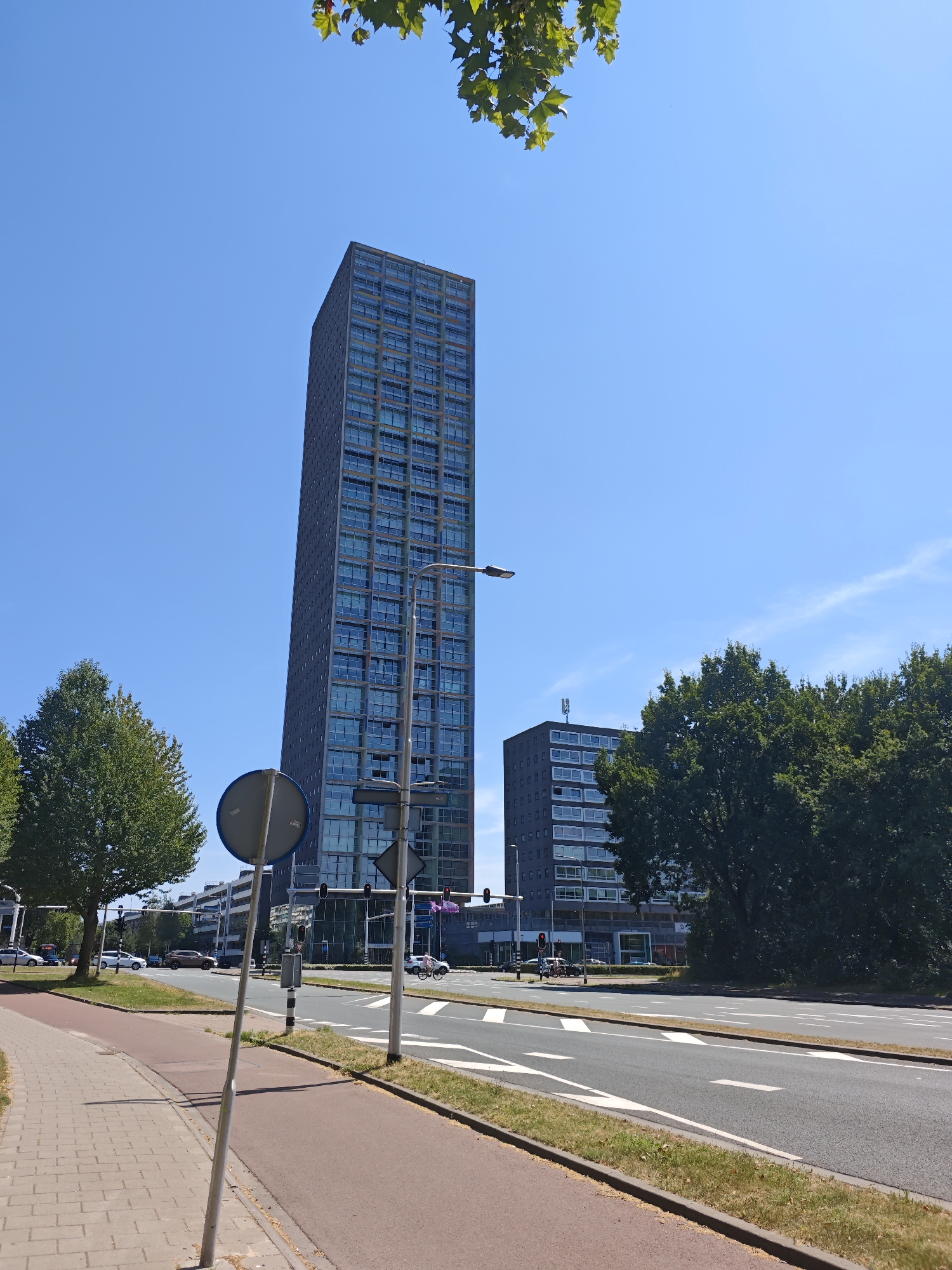 A bright, sunny day in Tilburg, Netherlands, features a tall, modern skyscraper with numerous windows, standing beside a lower, gray building. In the foreground, a paved sidewalk and a red bicycle path run alongside a multi-lane road. Several cars are visible on the road, and traffic lights are present. Lush green trees line the street and partially obscure the buildings. The sky is a clear, vibrant blue, with a few wispy clouds. A metal pole with a circular sign is positioned on the sidewalk.