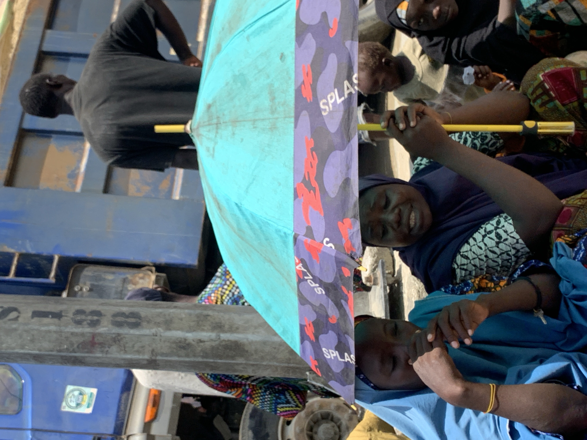 This outdoor scene, likely captured in Lagos, Nigeria during bright daylight, is rotated 90 degrees clockwise. It features several people gathered around a large blue truck.
In the foreground, a group of women and children are visible. A woman on the left (when viewed upright) smiles warmly, holding the yellow handle of a partially opened umbrella. The umbrella is distinctive, with one half light blue and the other a purple camouflage pattern adorned with red shapes and the word "SPLAS" repeated in white text. Another woman, dressed in blue, is partially visible to the right, appearing relaxed. Multiple children's faces can be seen in the background, some looking towards the camera, others elsewhere. They are dressed in various patterns and colors.
The main backdrop is a large blue truck. A man in a dark t-shirt stands on the truck bed in the background, facing away from the camera. On the side of the truck, faded black characters that resemble "T3S 88" are discernible, along with a small, unreadable circular sticker on the truck's cab. The bright sun casts clear shadows, indicating warm and sunny weather, with the umbrella providing shade for those beneath it. The overall mood appears calm and communal.