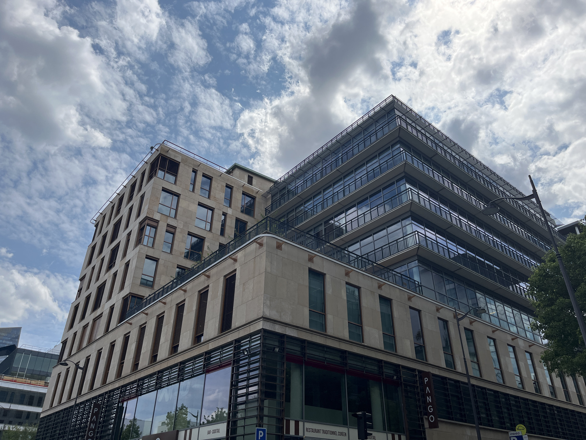 This is a low-angle shot of a modern, multi-story building against a partly cloudy sky. The building is a prominent subject, featuring a combination of stone and glass facades. The left side of the building is composed of light-colored stone with numerous rectangular windows. The right side is characterized by floors with extensive glass windows and balconies with dark railings, creating a more transparent and contemporary aesthetic.
The sky above is a mix of blue sky and scattered, fluffy white and gray clouds, suggesting a dynamic weather condition, possibly mid-day or afternoon. The lighting appears natural, casting some shadows but not indicating an extreme time of day.
Several text elements are visible on the lower levels of the building. On the right side, a vertical sign reads "Pango". Below the glass facade on the ground level, there are signs that are partially obscured but include text like "BAR - COCKTAIL" and "RESTAURANT TRADITIONNEL CORÉEN". A blue parking sign is also visible.
The scene depicts an urban setting with modern architecture. There are no people visible, and the focus is entirely on the building and the sky. The overall impression is one of architectural prominence and a typical day in a city environment.