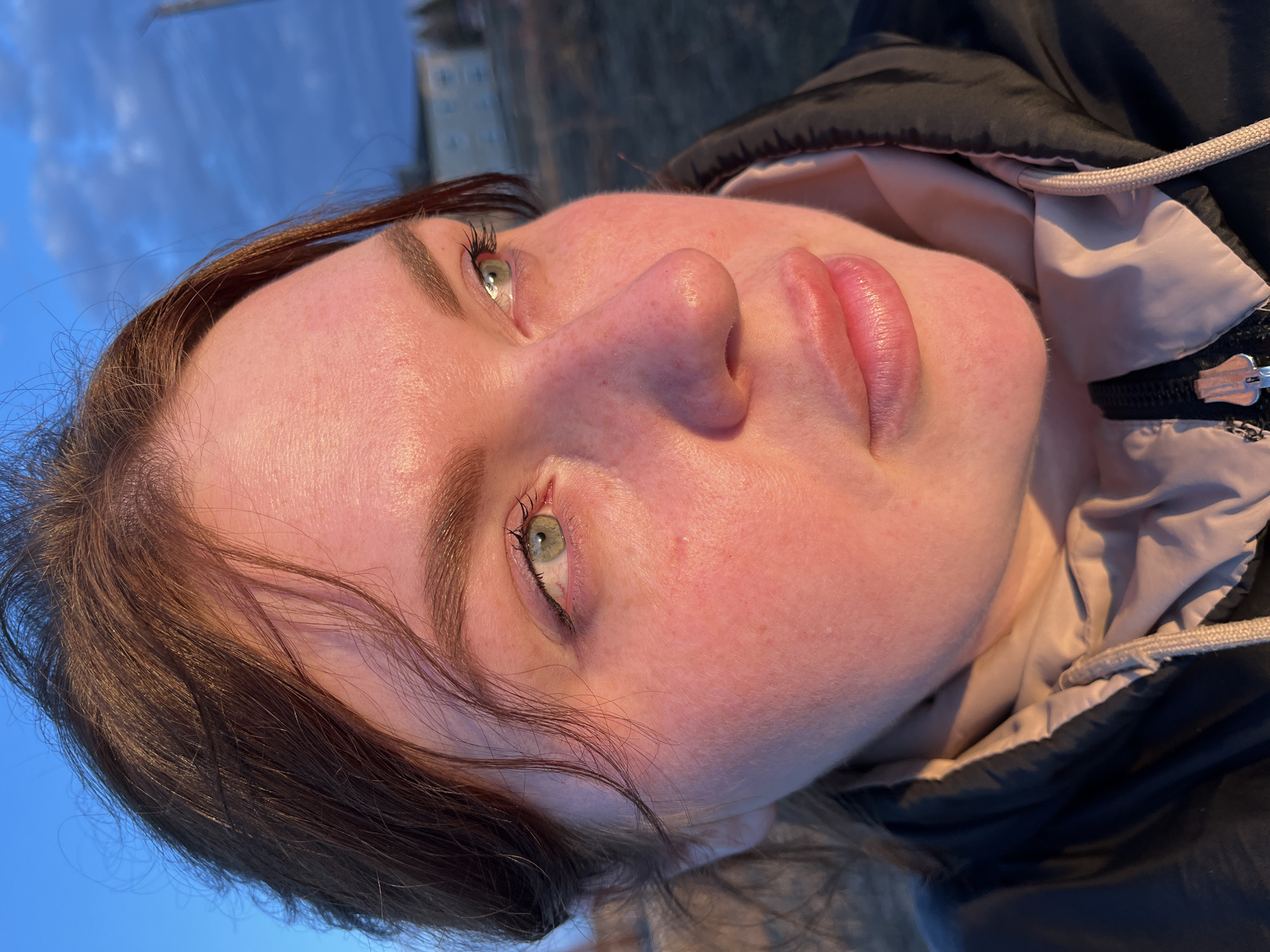A close-up, slightly rotated image shows a woman's face. She has fair skin, brown hair, and green eyes. She is wearing a black jacket with a light-colored lining. The background consists of a blurred cityscape under a blue sky, which could potentially locate the image in the city of Павловка, Russia. The focus is primarily on her facial features and the details of her clothing. The lighting suggests it is either early morning or late afternoon.