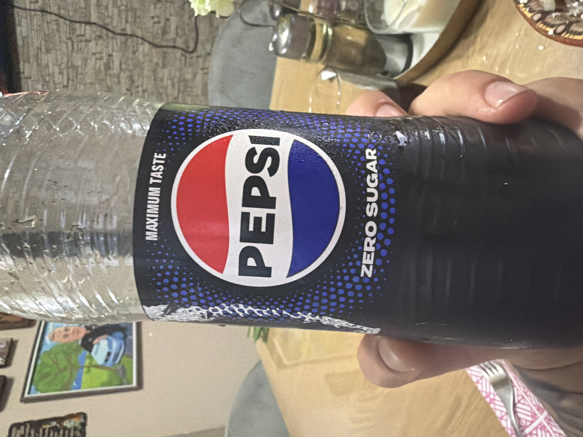 The image captures a close-up view of a plastic bottle of Pepsi Zero Sugar, held by a person's hand, in an indoor setting in Shumen, Bulgaria. The bottle is clear at the top, showing condensation and a ribbed texture, indicating it contains a cold beverage. Its dark blue/black label features the prominent red, white, and blue circular Pepsi logo. Vertical white text on the left of the logo reads "MAXIMUM TASTE," and on the right, "ZERO SUGAR." The background of the label is adorned with a pattern of small blue dots.
A hand with light to medium skin tone is gripping the bottle. The immediate background shows a wooden table surface. Various household items are visible, including reflective glassware or condiment bottles, and a soft, white floral arrangement in the upper left. A wall with textured, possibly wallpapered, surface in a grey tone forms the backdrop. On this wall, a framed picture depicting a person in a green top and blue headgear is visible in the lower left, with another smaller framed object below it. A silver fork is resting on a pink and white patterned napkin in the lower right corner of the frame. The scene suggests a casual indoor setting, likely a dining area.
