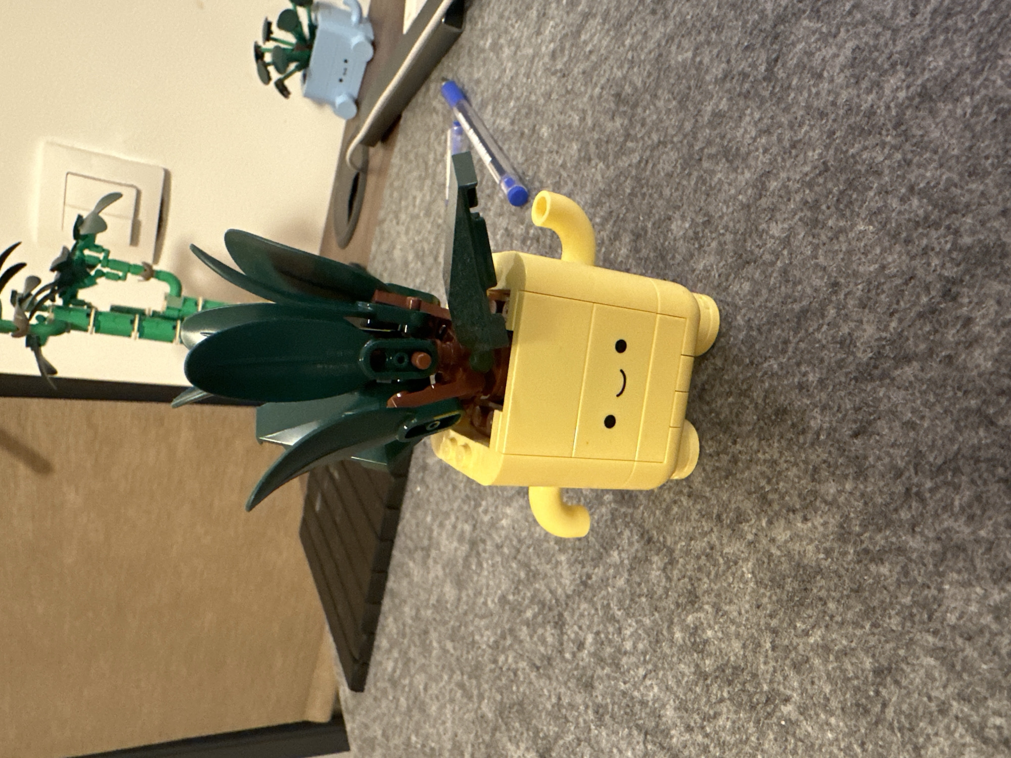 A close-up, high-angle shot reveals a yellow, square-shaped Lego head of a pineapple with an inserted pineapple stem, sitting on a gray, textured surface. The head features a simplistic smiley face. Adjacent to the pineapple is a green Lego cactus. A blue pen lays nearby. The background includes a light-colored wall with an electrical outlet and part of a brown cabinet. The location is not discernible from the image.