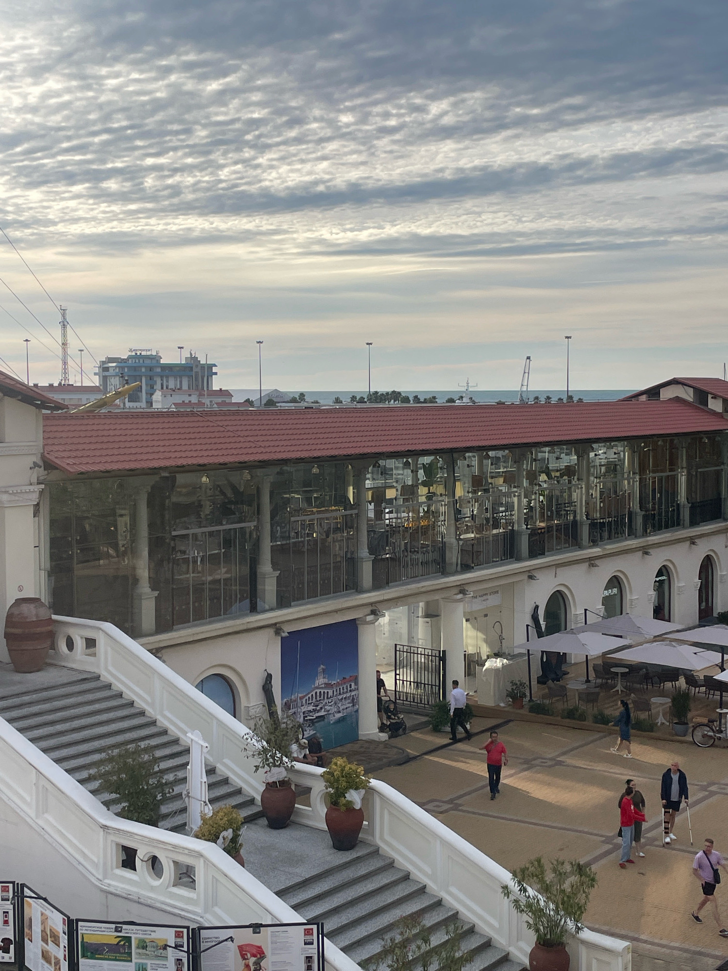 The image captures an outdoor area in Sochi, Russia, featuring a multi-story building with a red-tiled roof and large glass windows. The building appears to be a restaurant or cafe, with tables and chairs visible both inside and outside under umbrellas. A grand white staircase with ornate railings leads down from the upper level, adorned with large potted plants. Several people are walking around the plaza area in front of the building, some are sitting at tables, and one person is leaning on a crutch. In the background, a body of water, possibly the sea, is visible with ships and cranes in the distance, indicating a port or harbor. The sky is filled with dramatic, textured clouds. The overall scene suggests a public space in a coastal city.