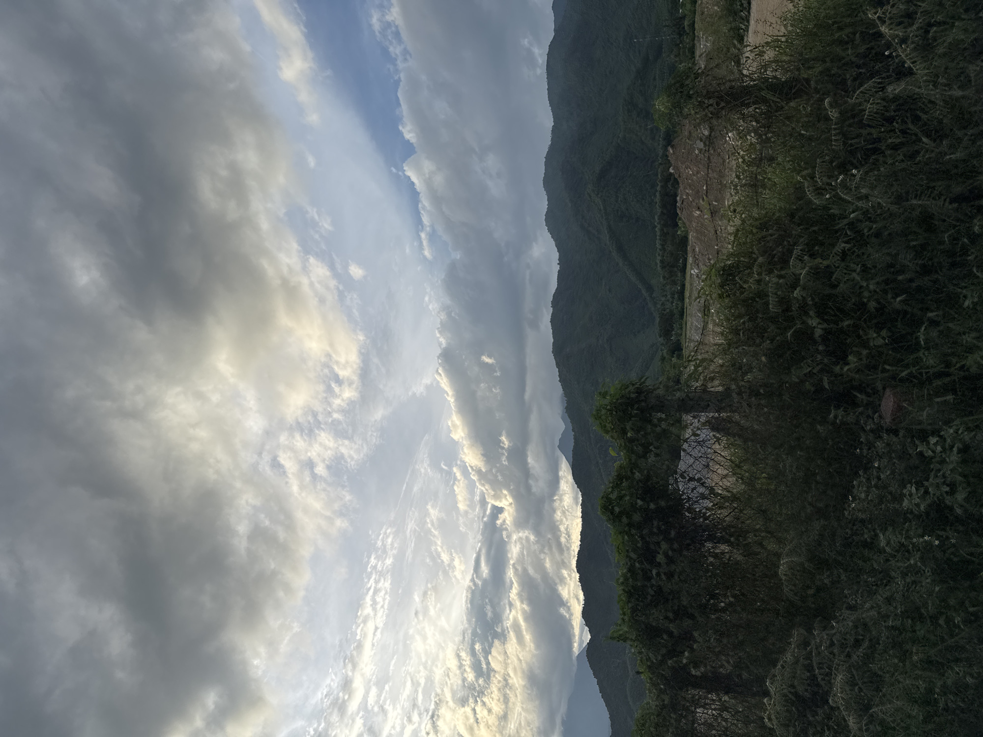 The image depicts a cloudy sky over a mountainous landscape. In the foreground, dense green foliage and a chain-link fence covered in vines are visible. Beyond the foliage, a dirt road or construction area with a pile of soil is present. The background features rolling green mountains under a dramatic sky with patches of sunlight breaking through the clouds. This scene is characteristic of the outskirts of Da Nang, Vietnam, likely in a rural or developing area. No people or specific actions are depicted.
