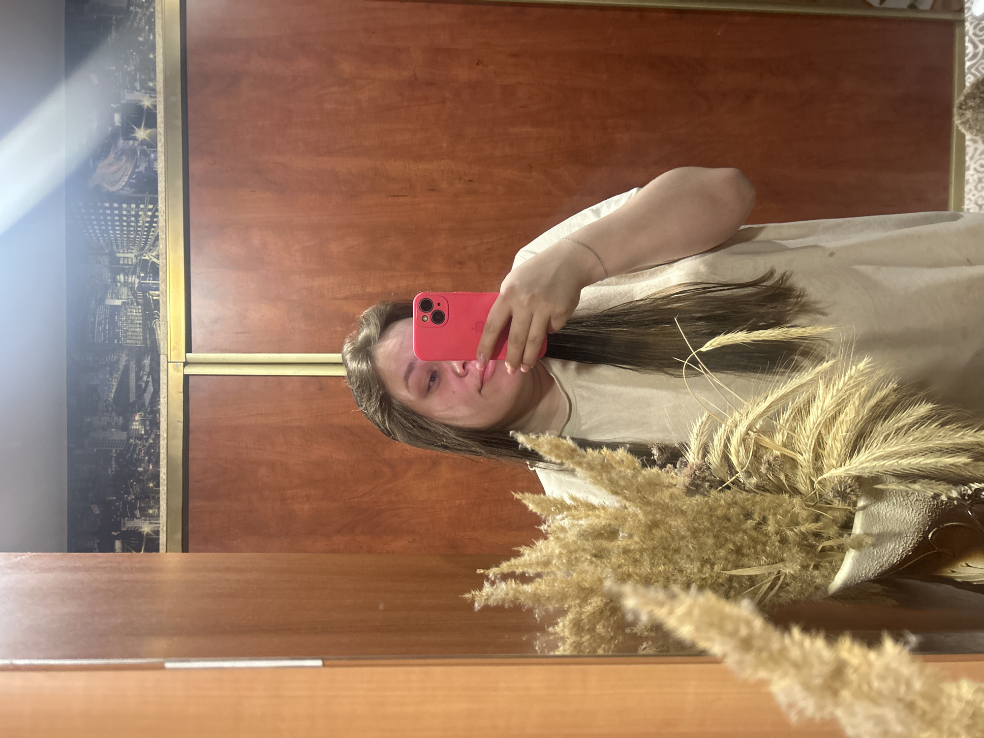 A woman is taking a selfie in a mirror. She is holding a pink cell phone. Her hair is long and brown. She is wearing a light-colored t-shirt and a silver bracelet. There is a bouquet of dried flowers in the foreground. The background is a wooden wall. On the left side of the wall is an image of a city skyline, possibly New York. The mirror is framed with wood. There is no definitive information to suggest that this image was taken in Ольховка, Russia.
