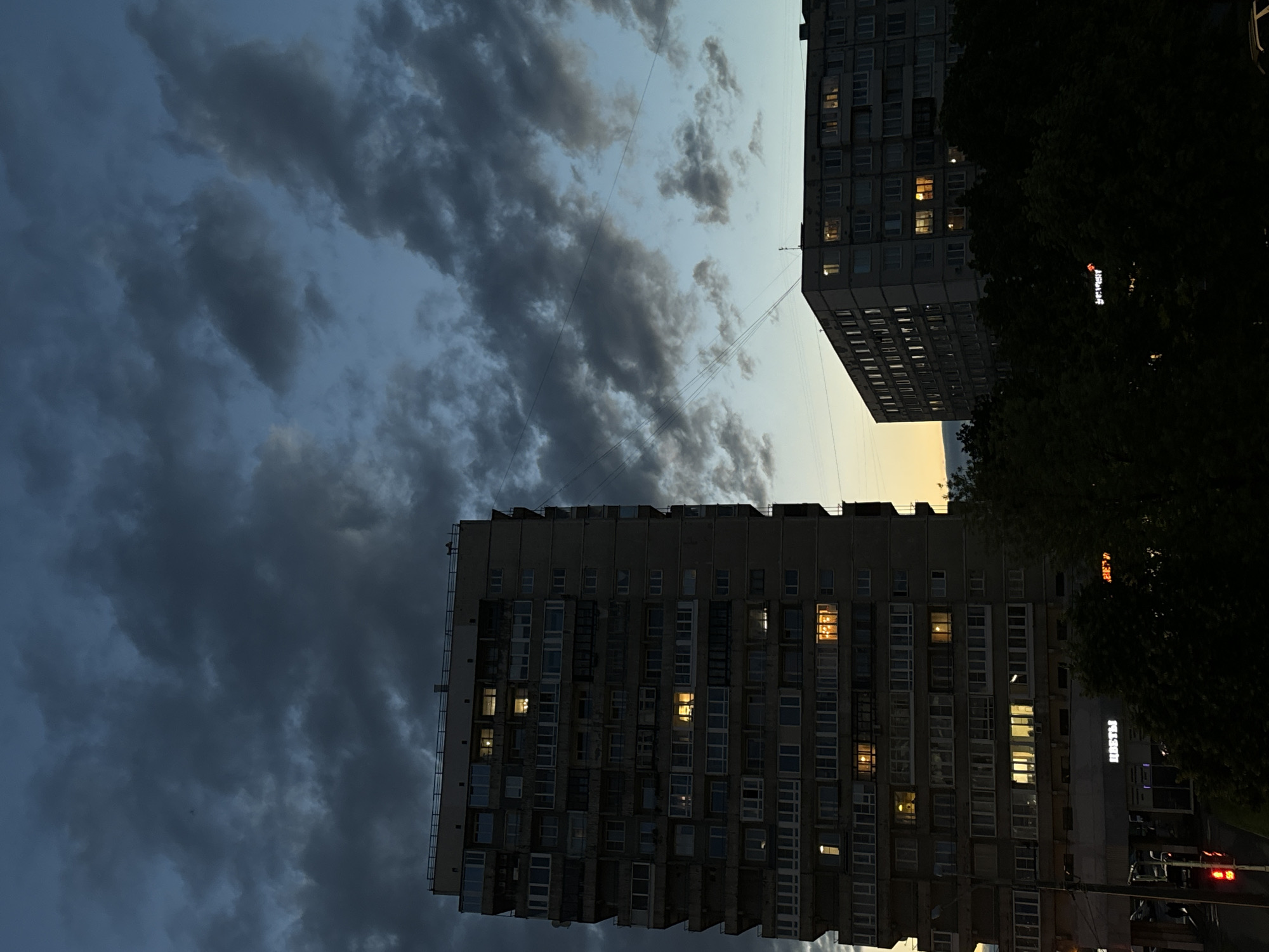 The image shows a view of two apartment buildings in Saint Petersburg, Russia, at dusk. The sky is filled with dark, dramatic clouds, transitioning from a deep blue at the top to a lighter, yellowish hue near the horizon. The taller building on the left is a multi-story structure with numerous windows, many of which are illuminated with a warm, yellow light, suggesting that people are inside. Power lines are visible crisscrossing the sky. To the right, a shorter, wider apartment building is also visible, with some illuminated windows. Trees, silhouetted by the fading light, are in the foreground, obscuring the lower parts of the buildings and the street level. One of the buildings has a sign that reads "ЦВЕТЫ," which translates to "FLOWERS" in English, indicating a flower shop or business on the ground floor. The overall scene conveys a sense of evening settling in over an urban environment.