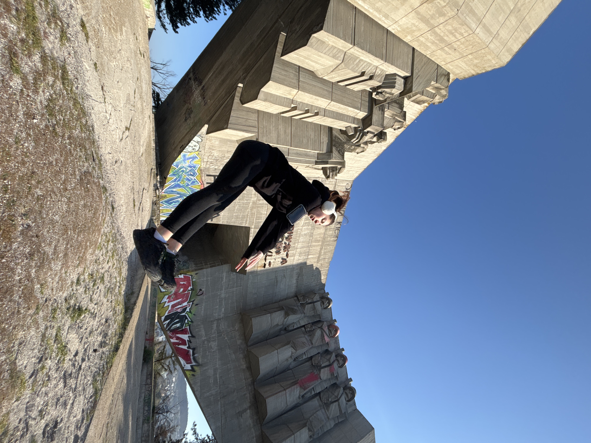 A full, angled shot of a woman in black performing a yoga pose on top of a concrete structure in an urban environment. The woman is wearing black leggings, a black top, black shoes, and white earmuffs. She is in a downward-facing dog pose, with her hands on the concrete structure in front of her and her feet on a section of the structure behind her. The concrete structure is covered in graffiti in some areas and features a series of concrete sculptures. The sky is clear blue. The location is in the city of Unknown, Unknown.