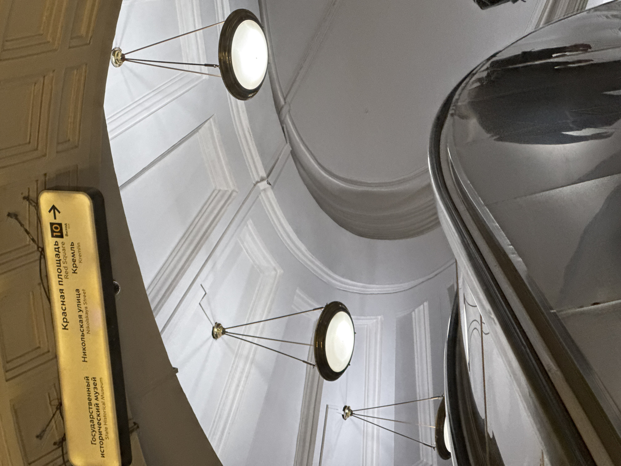 This is a medium-shot, low-angle photograph taken inside a Moscow Metro station, likely the "Okhotny Ryad" station, which serves as a hub connecting to the Red Square, Kremlin, Nikolskaya Street, and the State Historical Museum. The image captures an overhead directional sign with Russian and English text indicating these locations and an arrow pointing towards exit 10. The sign is illuminated and mounted on a decorative ceiling. Two circular, pendant light fixtures with brass-colored frames and white diffusers hang from the ceiling via thin wires, casting a warm glow. In the foreground, a portion of a highly polished, metallic escalator handrail curves into view, reflecting the ambient light and the ceiling architecture. The background features a white, ornate ceiling with recessed panels and molding, characteristic of Soviet-era metro station design. The overall impression is one of architectural grandeur and efficient transit signage.