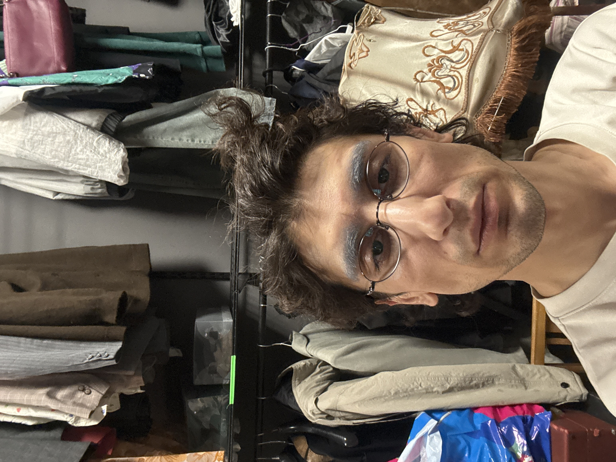 A man with dark, curly hair, glasses, and stubble is captured in a close-up selfie, looking directly at the camera with a neutral expression. He is wearing light-colored, possibly cream or beige, shirt and has prominent blue/grey eyeshadow on his eyelids.
The background reveals a densely packed indoor space, likely an open closet or shelving unit in a residence in Moscow, Russia. Black metal shelves are stacked with various folded clothes and personal items. On the left side, a maroon handbag sits atop a stack of clothes, including a green garment and light-colored fabrics. Behind the man's head, a decorative pillow or textile with an ornate golden-brown embroidered pattern and fringe is visible. To the right and below, more clothes are stacked, including a beige jacket and a vibrant blue, pink, and white patterned item, possibly a bag or fabric. A brown, suitcase-like object and a clear plastic box are also part of the stored items. A vertical green strip of tape or plastic is visible on the shelving unit. The image conveys an intimate glimpse into a personal, organized yet full, living space. There is no visible text. The time of day and external weather conditions are not discernible.