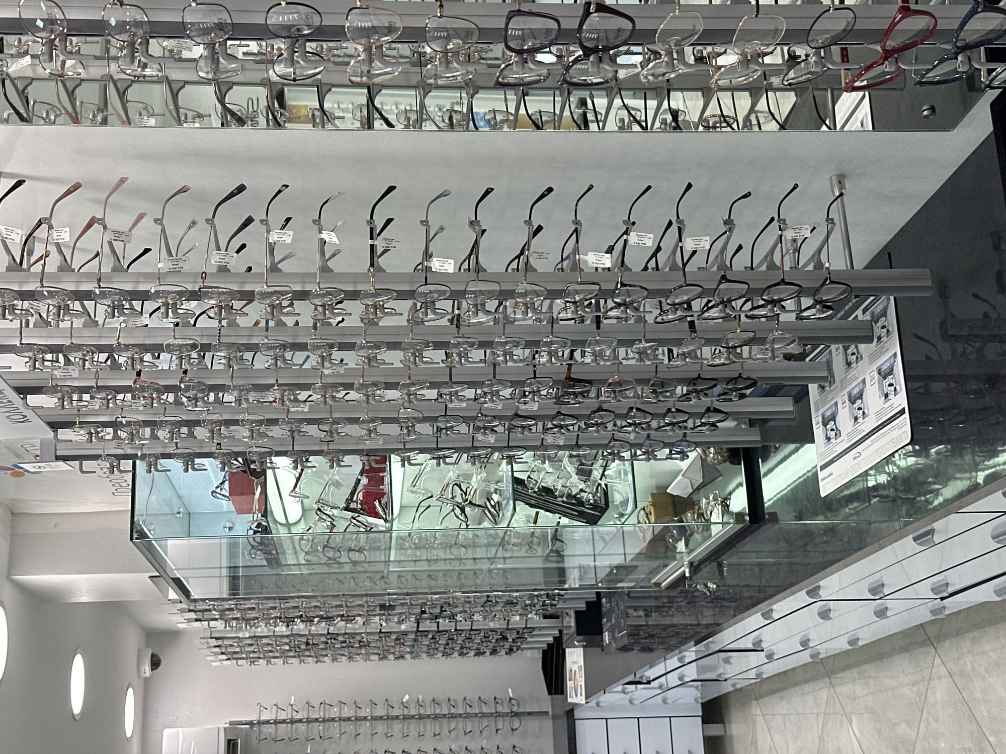The image shows the interior of an optical shop in Novoaltaysk, Russia. The shop is well-lit with recessed ceiling lights and features multiple displays of eyeglasses. On the left side, there are tall, narrow racks filled with eyeglasses, with additional displays visible in a glass-enclosed cabinet in the center. The right side of the image shows more vertical racks of eyeglasses, some with price tags. The foreground features a polished, dark countertop with drawers underneath, and a tiled floor. There are no people visible in the image. The overall impression is of a modern and organized retail environment.