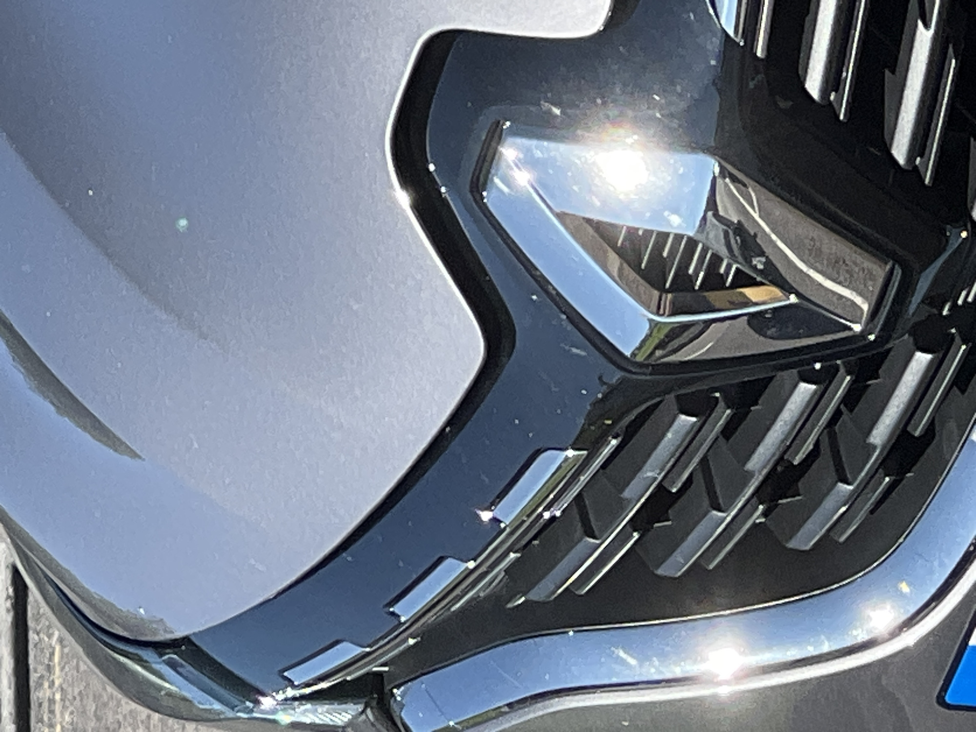The image shows a close-up view of the front grille and hood of a car, likely a Renault given the diamond-shaped emblem. The car is dark grey and appears to be parked outdoors, possibly in a city. Sunlight reflects off the car's chrome accents and the hood, creating some glare. The background shows a blurred asphalt surface, suggesting a road or parking lot. There is no indication of the city of Donegal, Ireland, in the image itself.