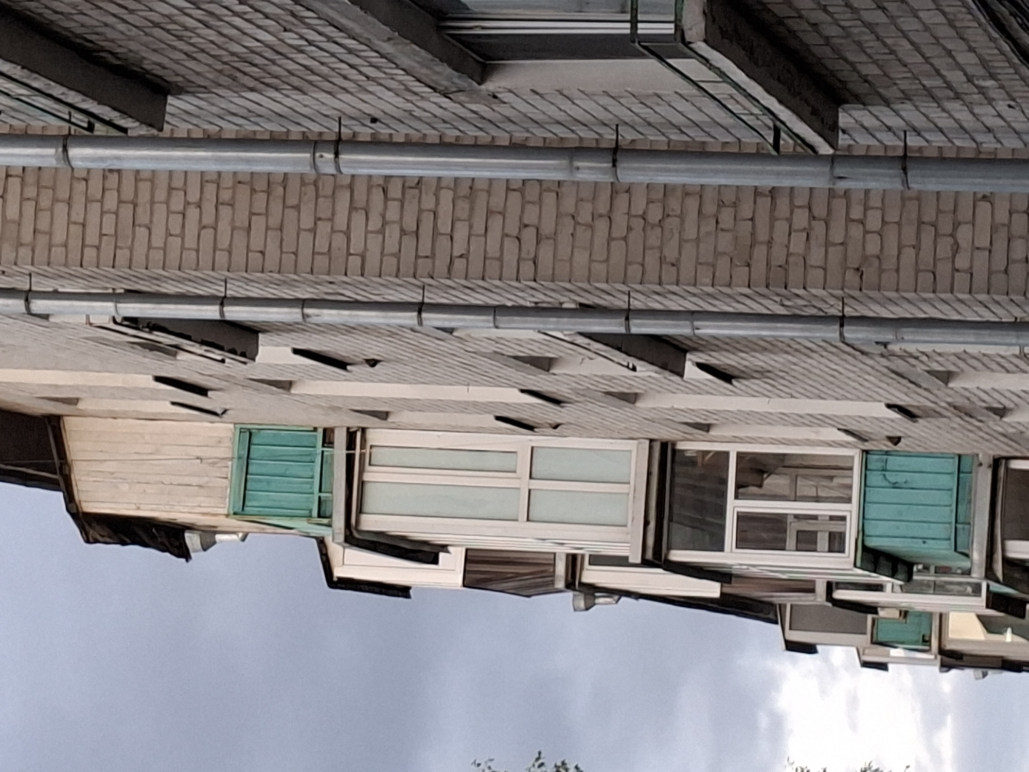 The image shows a close-up, low-angle view of the side of a multi-story brick building in Kazan, Russia. The building features multiple balconies, some enclosed with glass and white frames, and others with turquoise wooden shutters. Metal drainpipes run vertically down the exterior brick wall. The sky is overcast with gray clouds. The left side of the image shows a portion of another building or an extension with wooden siding and dark roofing material. The brickwork appears to be a light beige color. The overall impression is of an urban residential building under a cloudy sky.