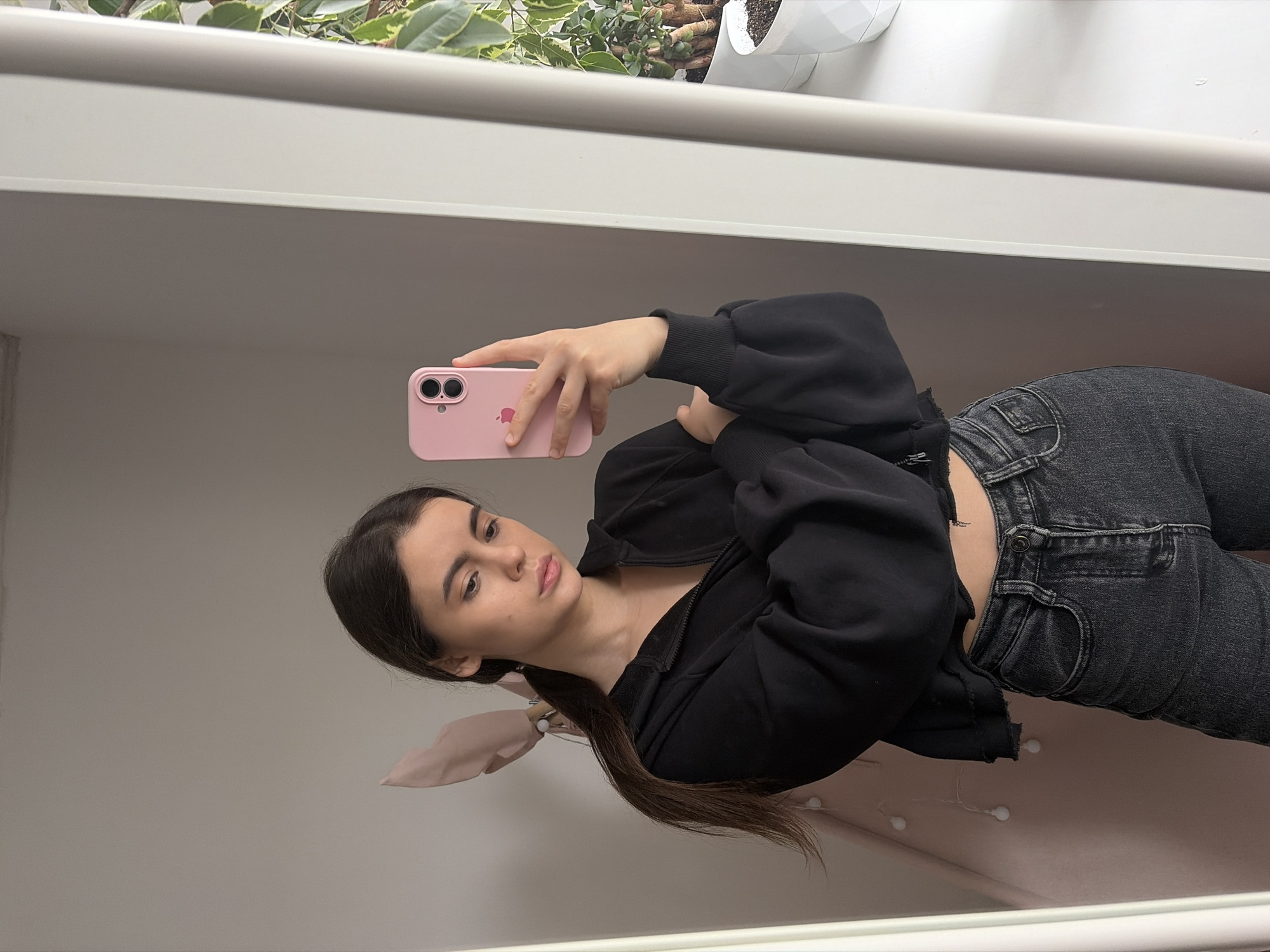The image captures a young woman, likely located in Chaikovsky, Russia, taking a mirror selfie indoors. She is positioned with her head tilted slightly, looking towards the camera of her smartphone. Her dark hair is pulled back into a ponytail, secured with a light pink or beige scarf-like fabric. She is dressed casually in a black zipped hoodie worn over a dark top, and dark gray jeans. Her expression is neutral and calm.
In her right hand, she holds a pink smartphone featuring a visible Apple logo on the back case and a dual-camera system. The background, which appears to be a mirror reflection, shows a light-colored shelf or beam running horizontally. Above this, there are glimpses of green leafy plants and a white pot, suggesting a domestic setting, possibly a living room or bedroom. The lighting in the scene appears soft and natural, indicating it is daytime. No other text is discernible within the frame.