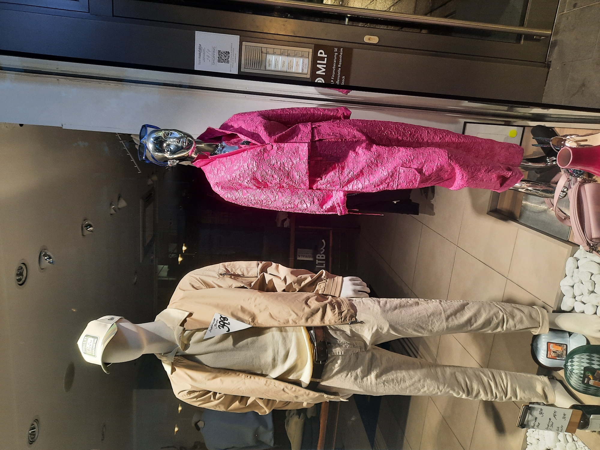 The image shows a storefront display window in Rosenheim, Germany. On the left, a male mannequin is dressed in a light beige bomber jacket, a light beige crewneck sweater, and light beige pants, topped with a light beige baseball cap. A price tag is attached to the jacket. On the right, a female mannequin is displayed wearing a bright pink textured jacket and matching pants, with blue sunglasses perched on its head. To the right of the mannequins, a sign with the MLP logo is visible, indicating it is an MLP Finanzberatung SE branch located in Rosenheim, Stock. The display features decorative white pebbles and a few handbags. The lighting suggests it is either daytime with the store lights on, or evening.