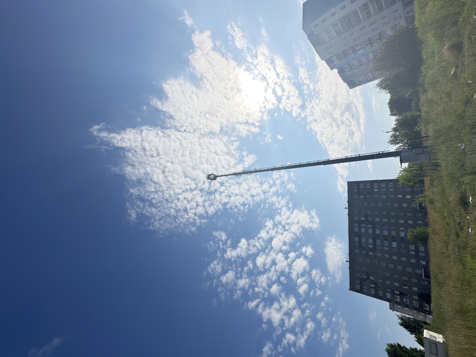 The media file is a photograph taken from a low angle, looking up at a bright blue sky filled with scattered white, puffy clouds. The sun is visible and slightly obscured by clouds, creating a bright glare.
In the sky, a long, slender object stretches diagonally across the frame. It appears to be a part of a crane or a tall structure, with a basket-like component at its extended end.
In the background, two multi-story buildings are visible on the right side of the frame. The building closer to the foreground is dark gray with numerous windows arranged in a grid pattern. The building further in the background is lighter in color and also features many windows. Between the buildings and the viewer, there is a strip of green grass and some trees, suggesting an outdoor setting. The overall impression is of an urban or suburban environment on a sunny day. The scene is likely Chaikovsky, Russia, based on the location context provided. The time of day appears to be daytime, with bright sunlight indicating midday or early afternoon. The weather seems fair, with clear skies and scattered clouds. There is no visible text in the image.
