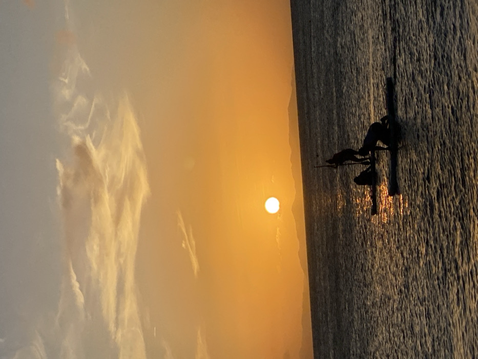 The image captures a sunset over a body of water in Juan Griego, Venezuela. The horizon is visible, separating a heavily orange and yellow-hued sky from the dark blue water. The sun, a bright, circular white-yellow orb, is located in the upper-left portion of the frame, positioned above the distant, silhouetted landmasses on the horizon. The sky exhibits scattered, wispy clouds.
Three individuals are present on the water, heavily silhouetted against the sunset reflections. One individual is kneeling on a paddleboard in the right foreground. A second individual stands upright on a paddleboard in the midground, holding a paddle vertically. A third individual is observed further to the left, positioned prone or very low on a paddleboard. The water's surface shows ripples and reflects the orange light from the setting sun. No other objects or specific interactions are discernable due to the heavy silhouetting.