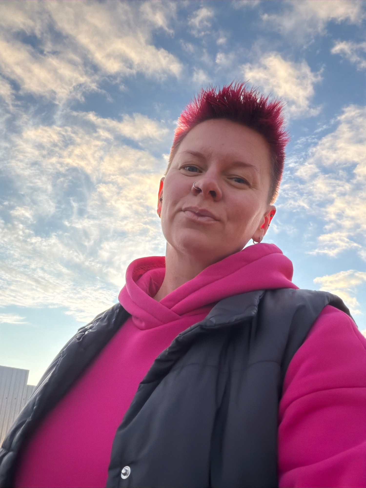 A low-angle, medium shot captures a person with bright pink, spiky hair, wearing a bright pink hooded sweatshirt and a dark gray vest, looking towards the camera with a neutral expression. The person has a nose ring and earrings. The background is a blue sky filled with scattered white clouds, with the sun partially visible behind the clouds, creating a bright glow. To the left of the frame, a portion of a light-colored building or structure is visible. The scene appears to be outdoors in a public space, possibly a street or an open area within the city of Chaikovsky, Russia, given the context.