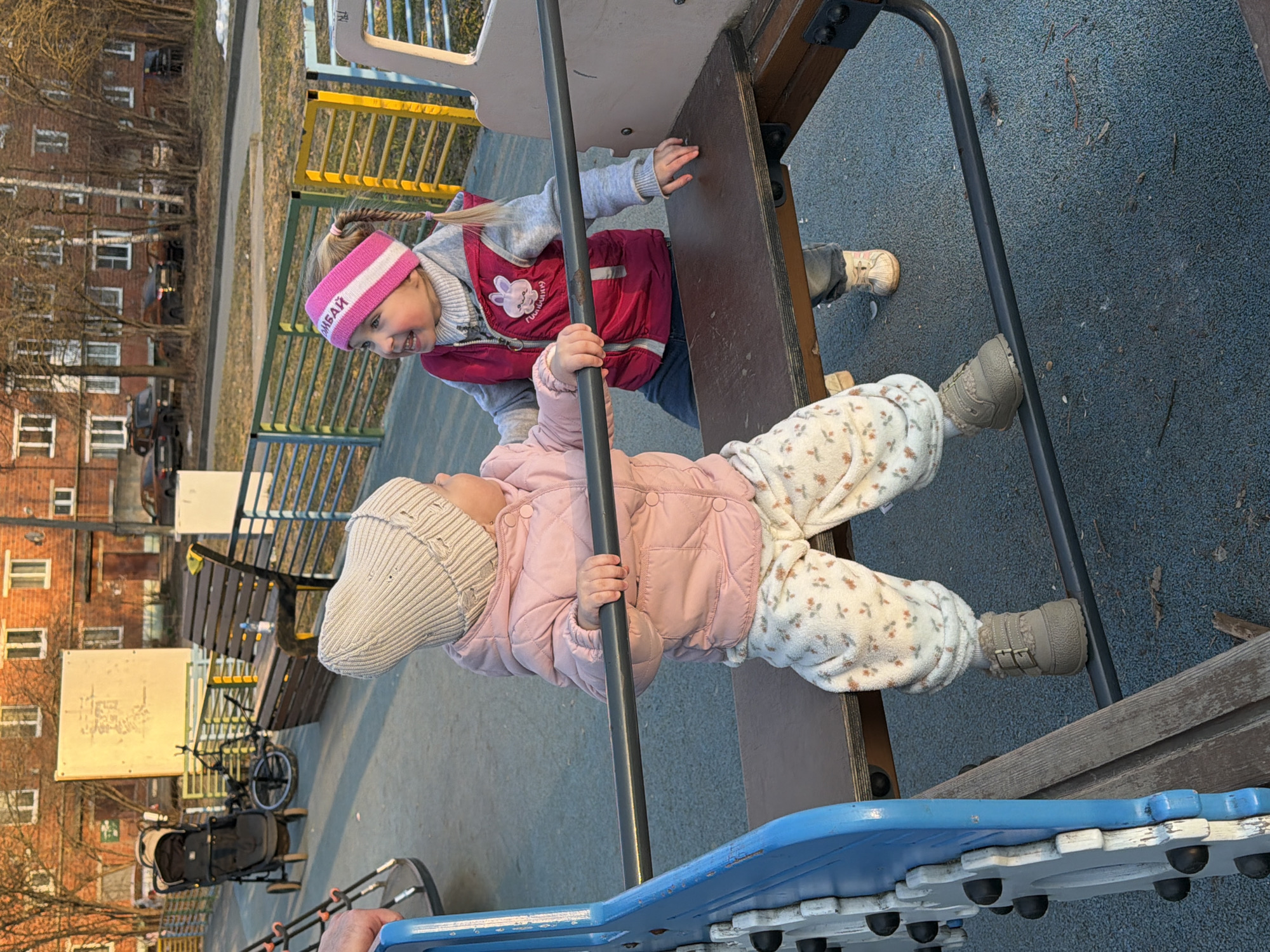 Two young children are playing on playground equipment in Reshetnikovo, Russia. The child on the left is wearing a pink coat, floral pants, a knit hat, and grey boots, and is holding onto a black metal pole. The other child, on the right, wears a pink headband, a pink vest, a grey sweater, and blue pants, and has a small ponytail. They are playing near a brown wooden structure, and there is a blue piece of playground equipment beneath them. A stroller and bike are visible in the background, along with a building and trees. The playground has a blue rubber surface.