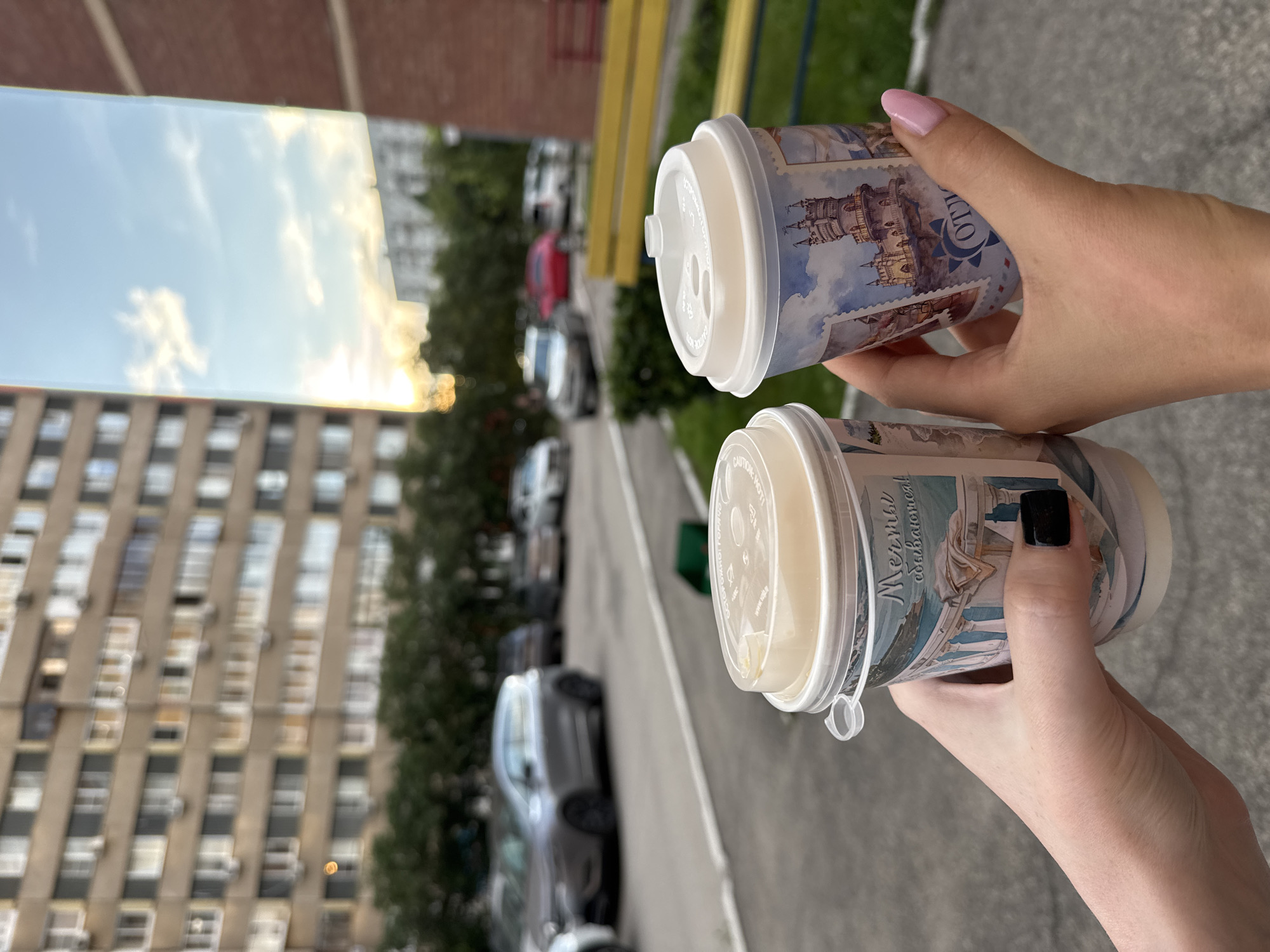 The image shows two disposable coffee cups being held by hands. The cups have decorative designs featuring castles and scenic views, printed in a style resembling postage stamps. The cup in the foreground has text that appears to be in Russian, reading "Мечты сбываются!" (Dreams come true!). The background is blurred but suggests an outdoor urban setting with a building, trees, parked cars, and a yellow fence. The sky is visible, reflecting in what appears to be a mirror or a reflective surface, showing clouds and a golden sunset or sunrise. The overall impression is a casual moment of enjoying drinks outdoors, possibly during a pleasant time of day due to the lighting.