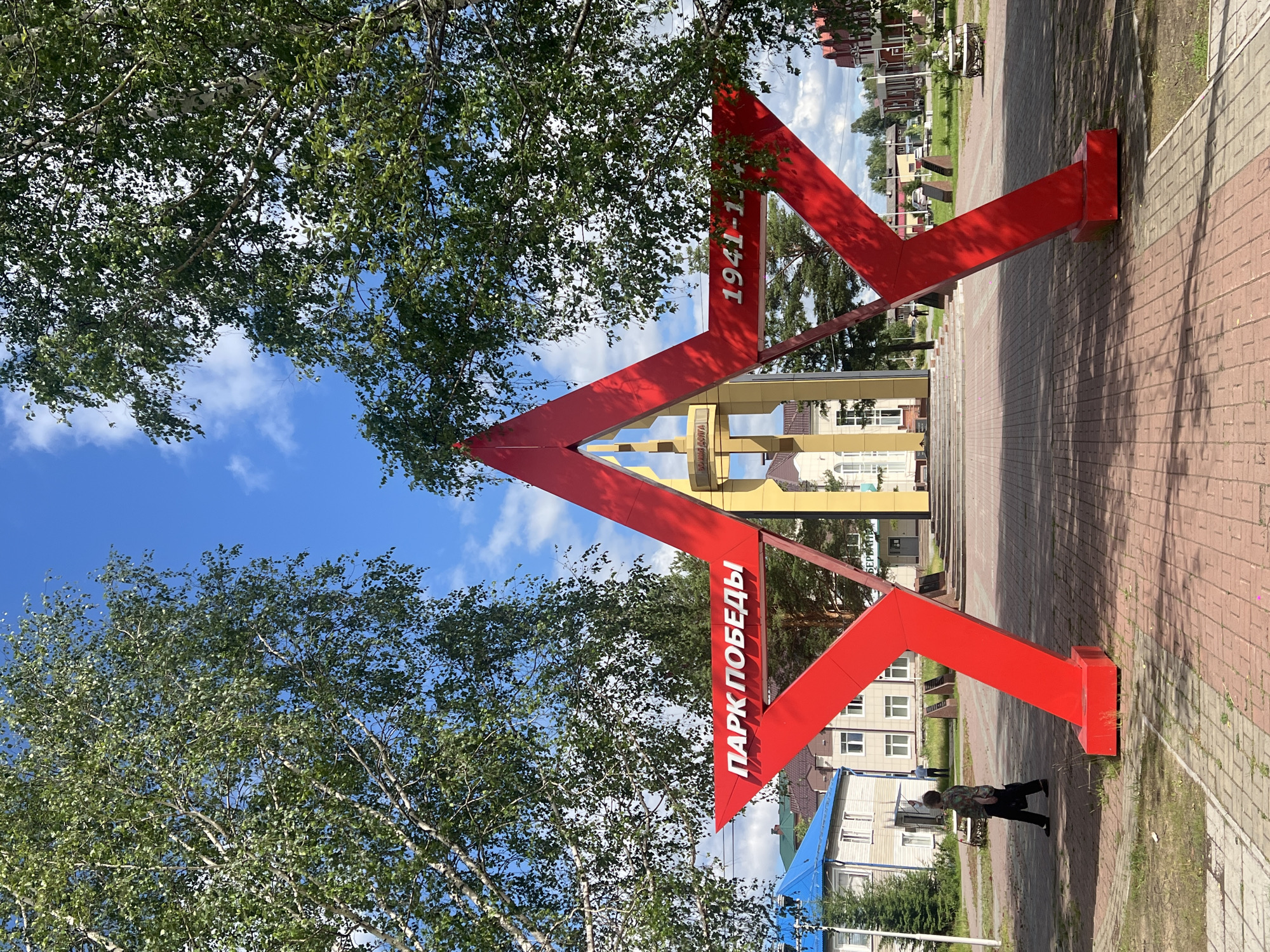 This is a vertical, eye-level, outdoor photograph taken in Mezhdurechenskiy, Russia. The image features a large, bright red, abstract sculpture with angular geometric shapes, set against a backdrop of trees with green foliage and a bright blue sky with scattered white clouds. The sculpture is in the foreground, dominating the center of the frame. It appears to be made of metal and has two distinct red angular elements that meet at a central point, forming an arrowhead-like shape.
Visible text on the sculpture reads "1941-1945" on one of its angled surfaces. On another surface, oriented vertically, the Cyrillic text reads "ПАРК ПОБЕДЫ" which translates to "Victory Park".
In the background, beyond the sculpture, there is a paved walkway or plaza made of red bricks. Further back, there are buildings with light-colored facades and pitched roofs, suggesting a residential or public area. The presence of trees and the blue sky with clouds indicate it is daytime, likely pleasant weather.
A small figure of a person can be seen in the lower right portion of the image, walking on the paved area. The overall impression is one of a public space, possibly a memorial or entrance to a park, marked by the prominent red sculpture commemorating a historical period. The bright colors and clear sky suggest a cheerful or celebratory atmosphere, despite the memorial inscription.