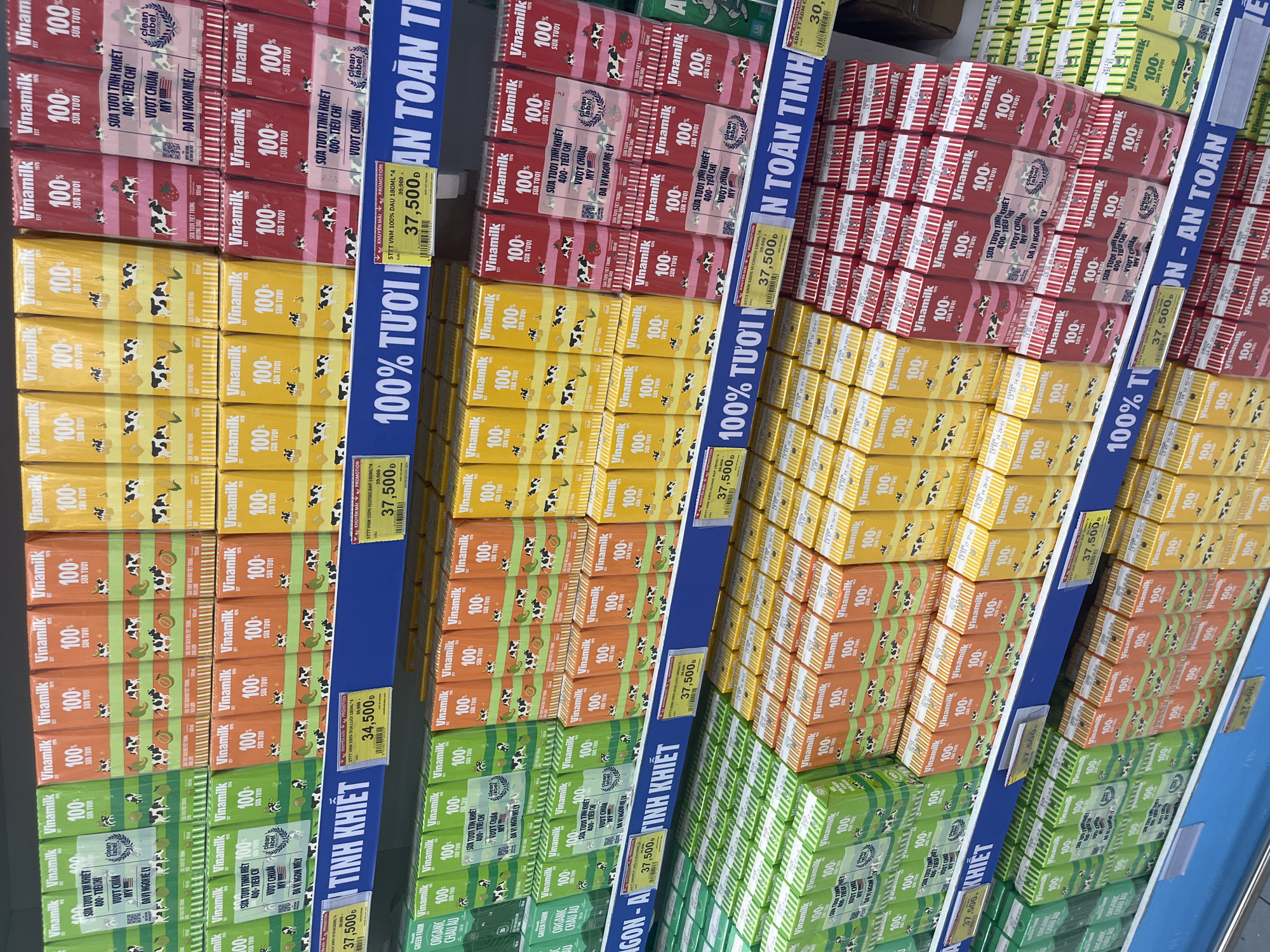 The image shows a retail display of VinaMilk 100% fresh milk cartons in various flavors, including green (durian), orange (melon), yellow (corn), and pink (strawberry). The cartons are neatly stacked on shelves in a supermarket. A blue shelf label in Vietnamese reads "100% TUOI" which translates to "100% Fresh" and "AN TOAN TINH KHIET" which translates to "Safe and Pure". Prices are displayed on yellow tags. The store appears to be well-lit and organized. The specific location is not visible in the image, but the branding and language suggest it is a supermarket in Vietnam.